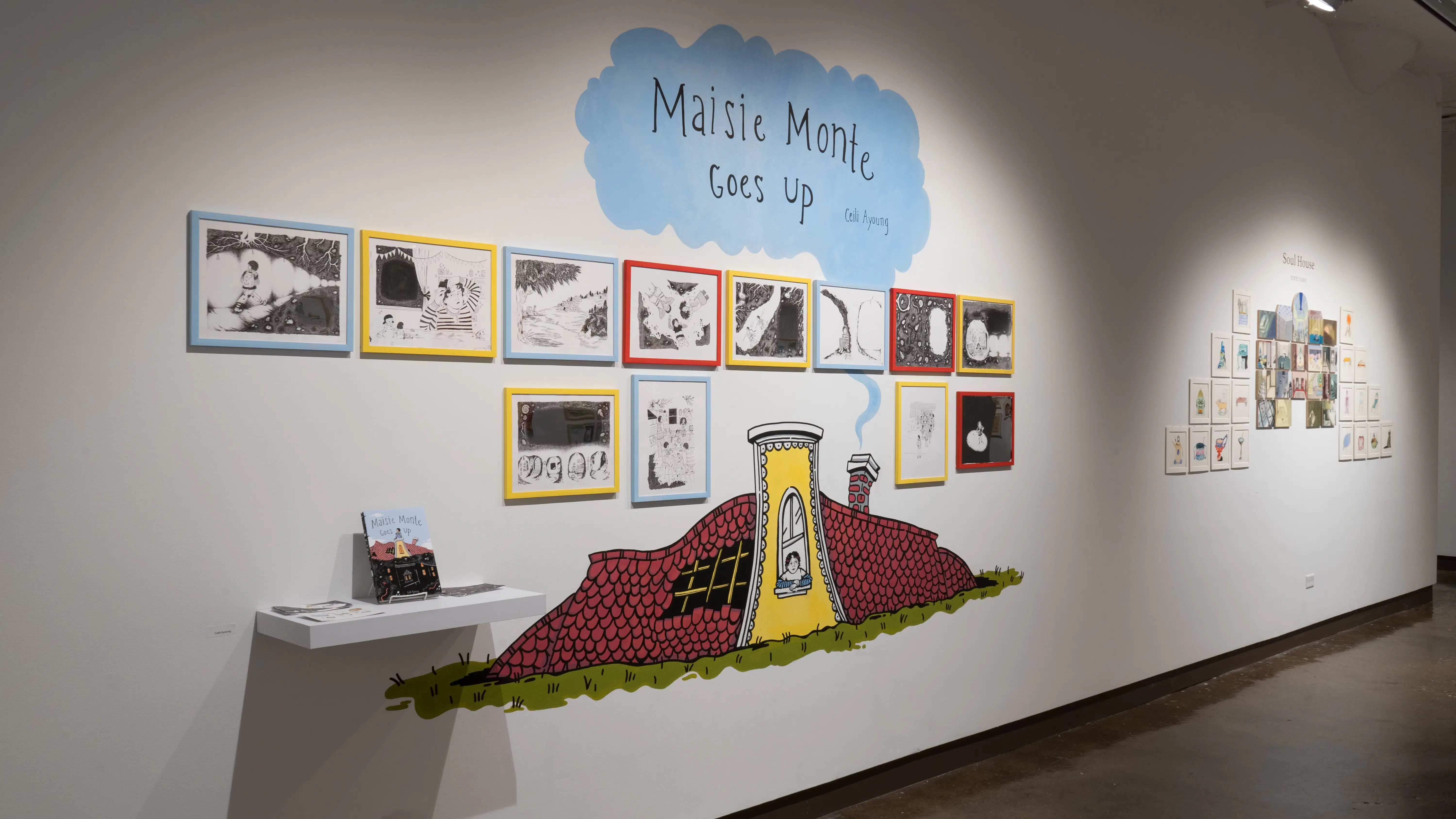 Black and white illustrations are framed in blue, yellow, and red frames and hung in two rows on a wall of gallery three. On the wall above the frames, “Maisie Monte Goes Up” is painted on a blue cloud, and below them, a small cottage is painted.