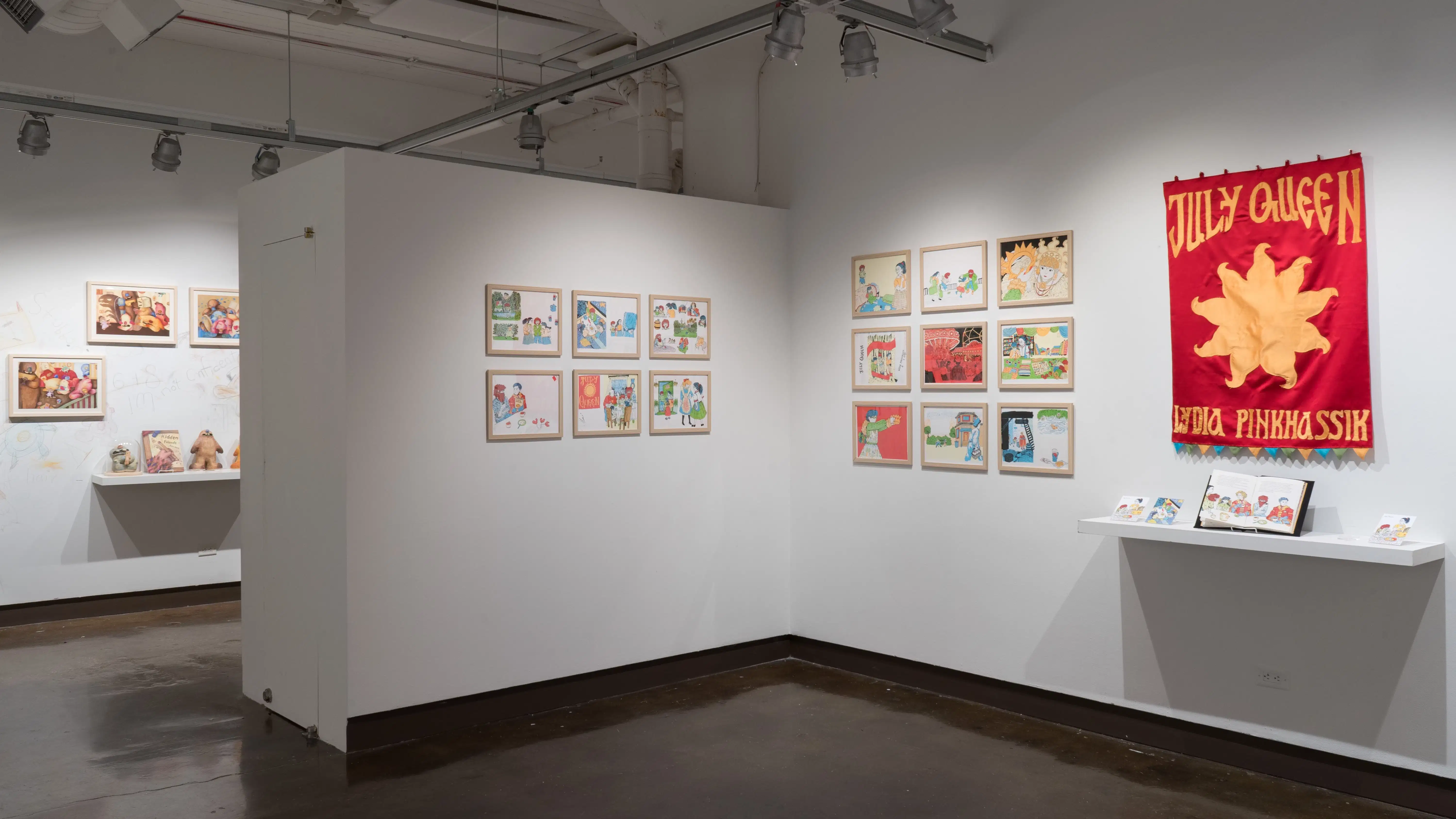 Illustrations in frames are arranged into grids in the gallery three alcove. In the center of the middle wall, a large red and yellow banner reads “July Queen - Lydia Pinkhassik.”