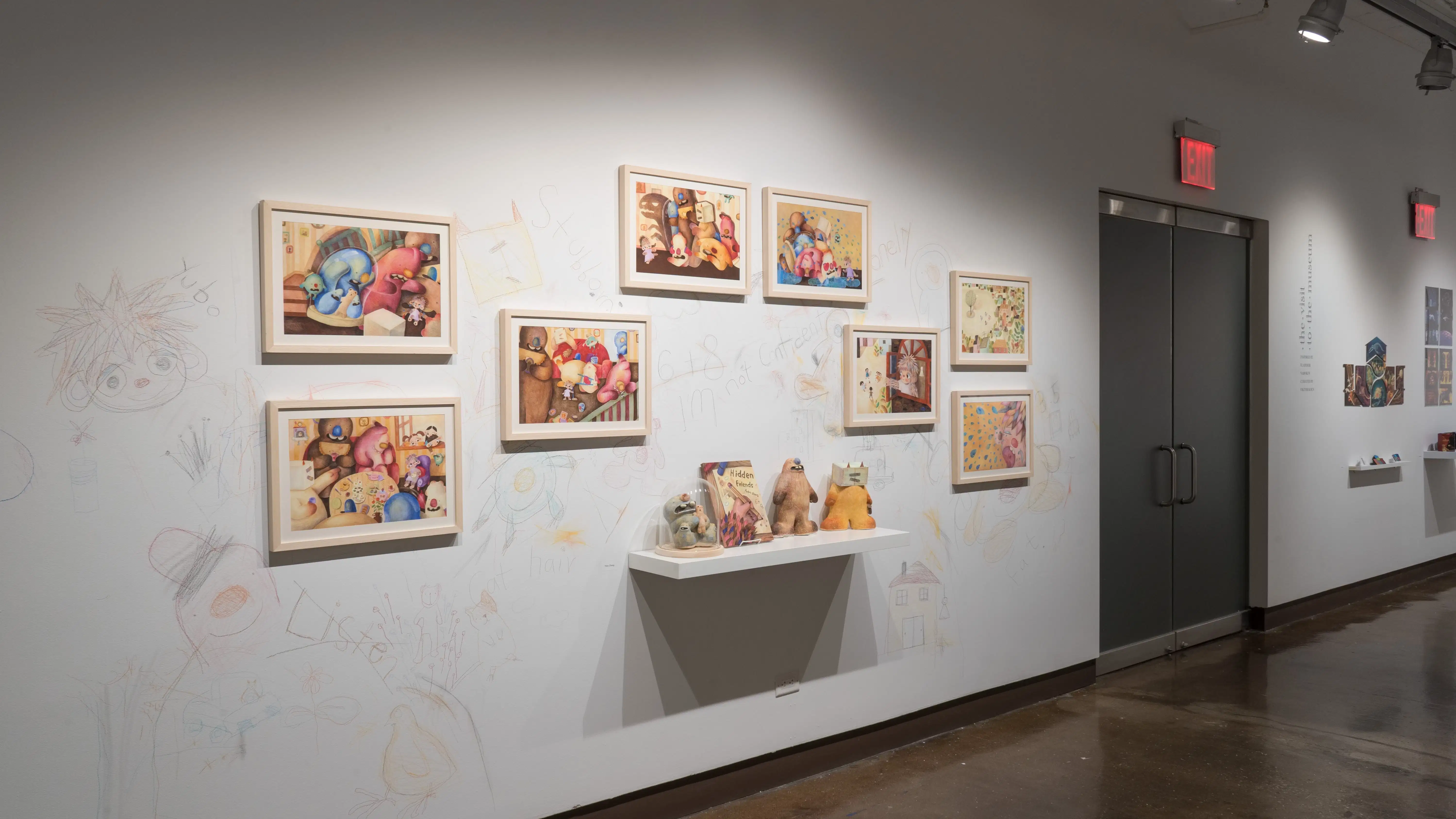 In the left hallway leading from gallery three to gallery four, colorful, framed illustrations are hung on the left wall. A shelf below them holds sculptures of the illustrated characters and a book. The surrounding wall is sketched on with colored pencils.