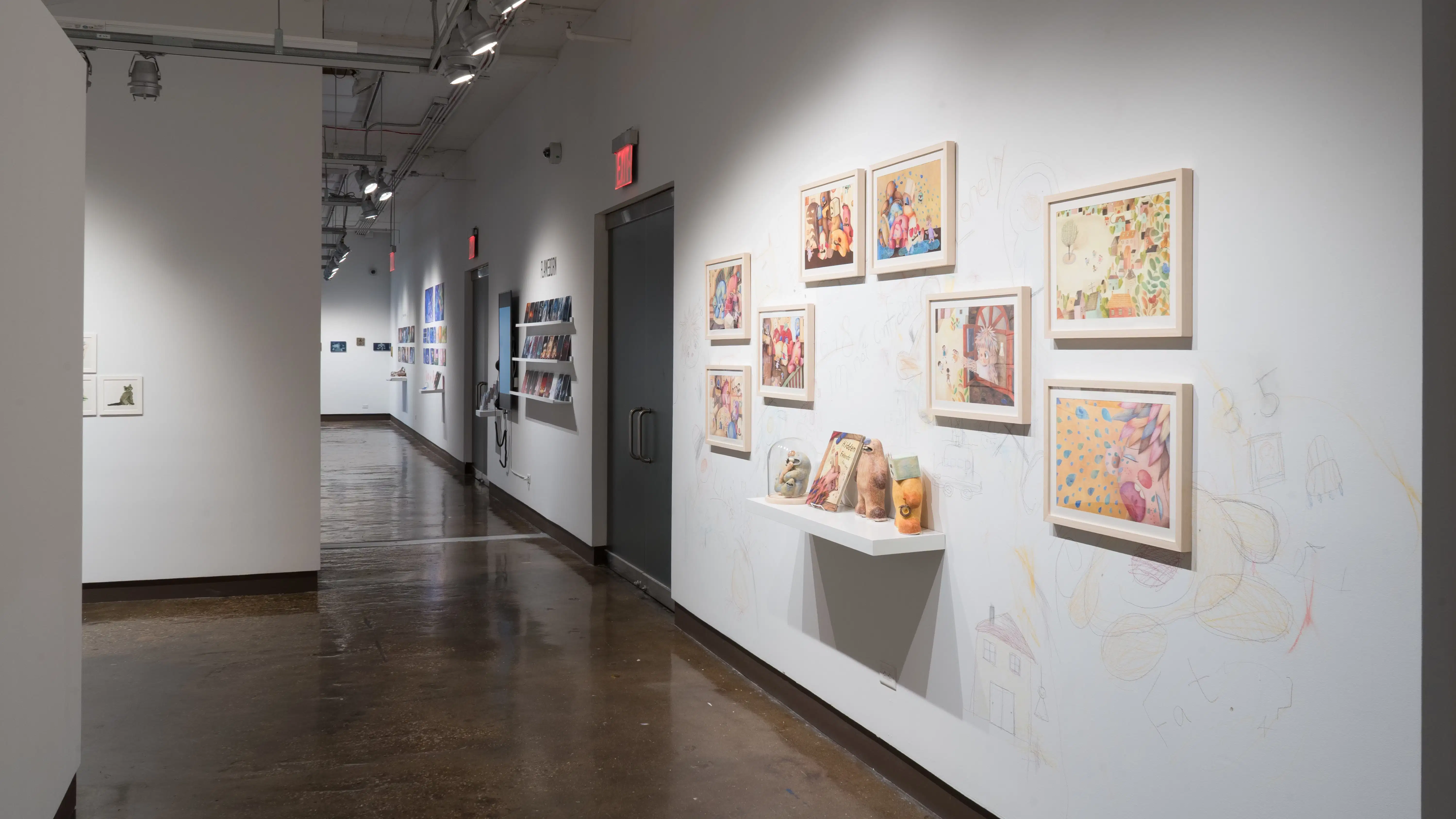 In the left hallway leading from gallery three to gallery four, colorful, framed illustrations are hung on the left wall. A shelf below them holds sculptures of the illustrated characters and a book. The surrounding wall is sketched on with colored pencils.