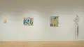 On one gallery wall, there is a pink and yellow painting on a small, squiggle-shaped panel. To the right, there are two figurative paintings in the same style. On the far right, a sculpture on the wall appears to be a long string of hair tied to a piece of metal covered with pantyhose.