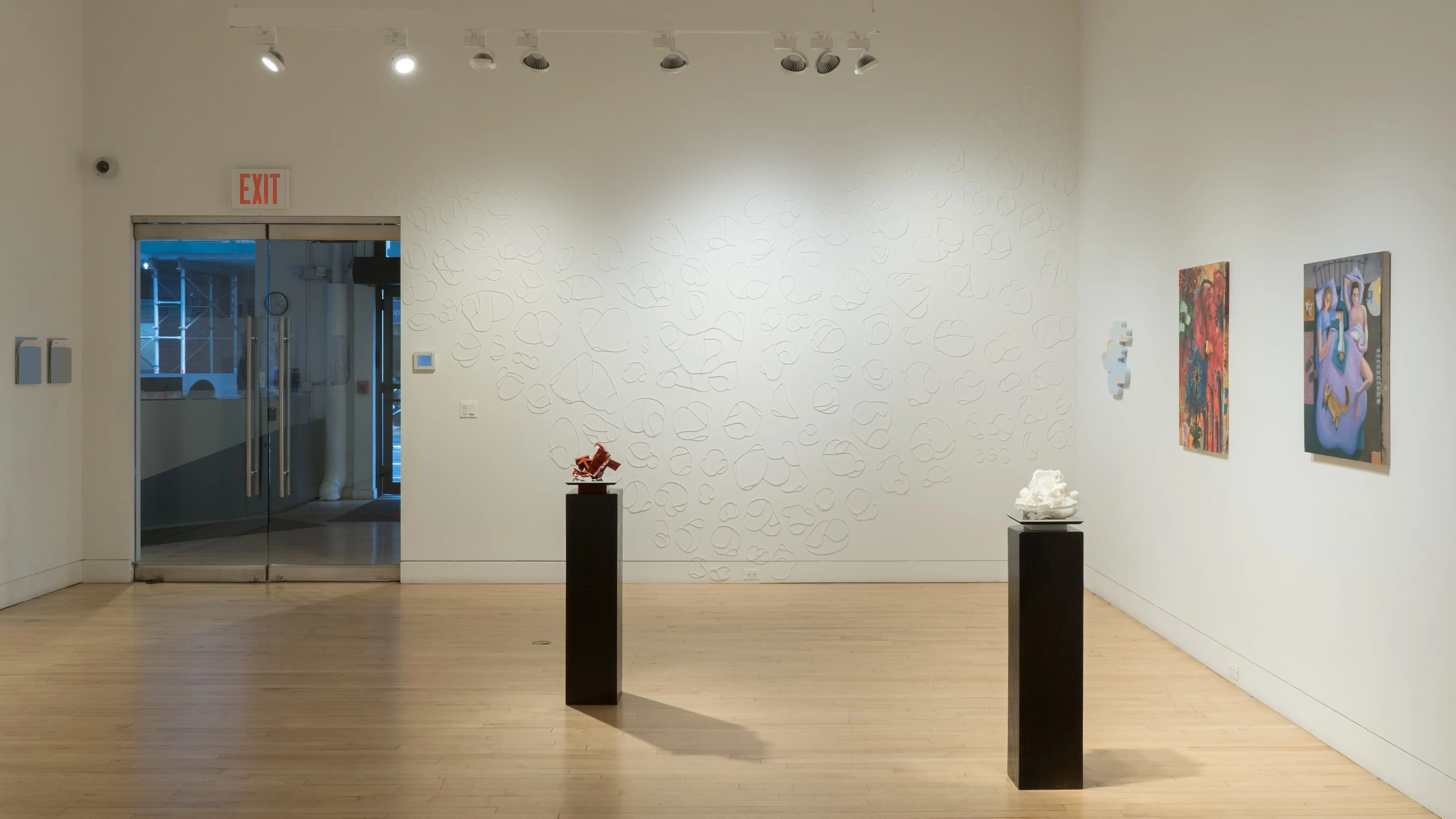In the center of the gallery, a red sculpture and a white sculpture are placed on their own black pedestals. The wall to the right holds three paintings, and the wall straight ahead is covered with zipties.