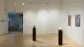 In the center of the gallery, a red sculpture and a white sculpture are placed on their own black pedestals. The wall to the right holds three paintings, and the wall straight ahead is covered with zipties.