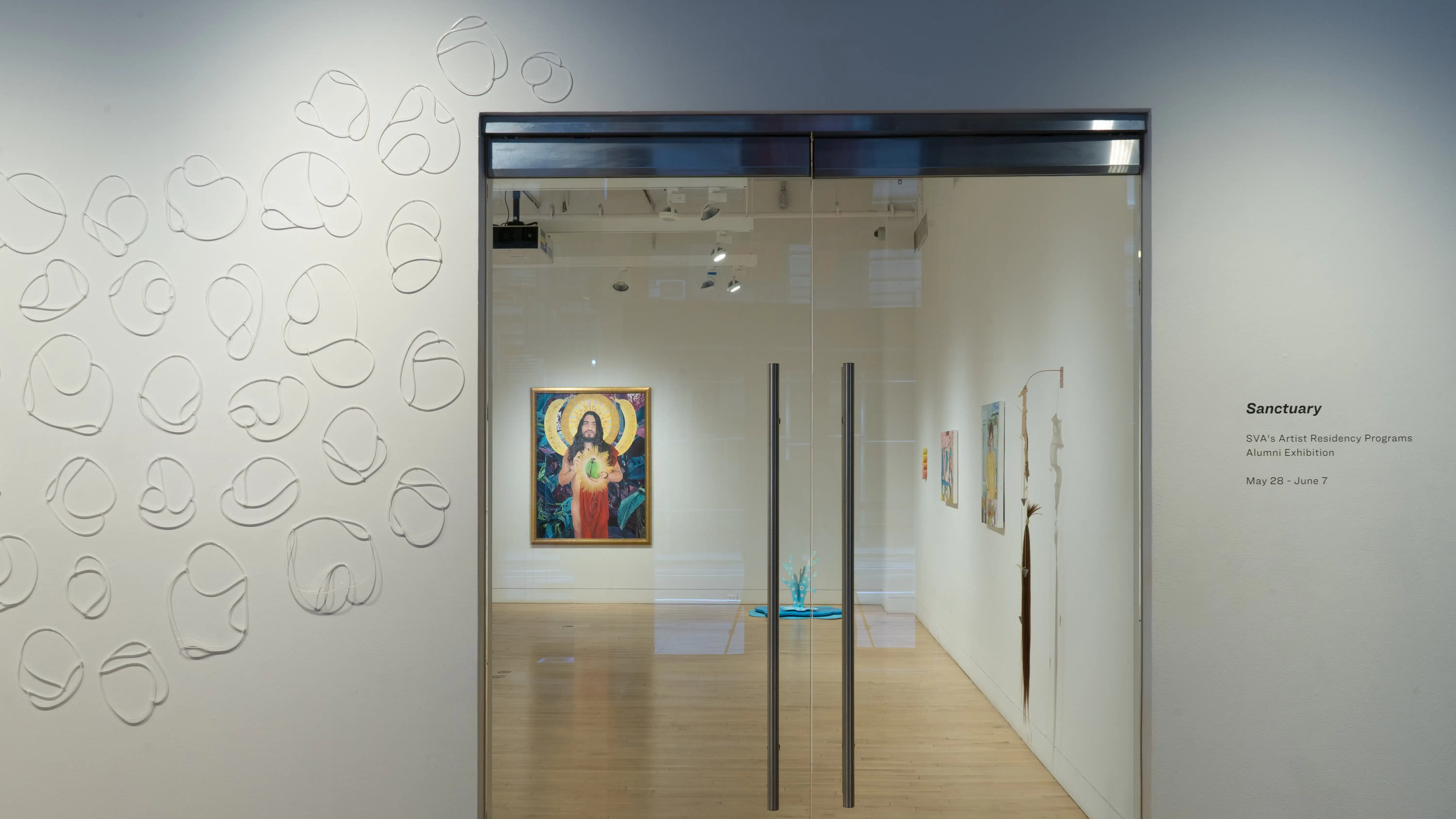 Facing the glass doors looking into the gallery, a painting of a saint or other holy figure hangs on the back wall. Sculptures and other paintings are also visible. Outside the door, zip ties are placed in a design on the wall.