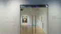 Facing the glass doors looking into the gallery, a painting of a saint or other holy figure hangs on the back wall. Sculptures and other paintings are also visible. Outside the door, zip ties are placed in a design on the wall.