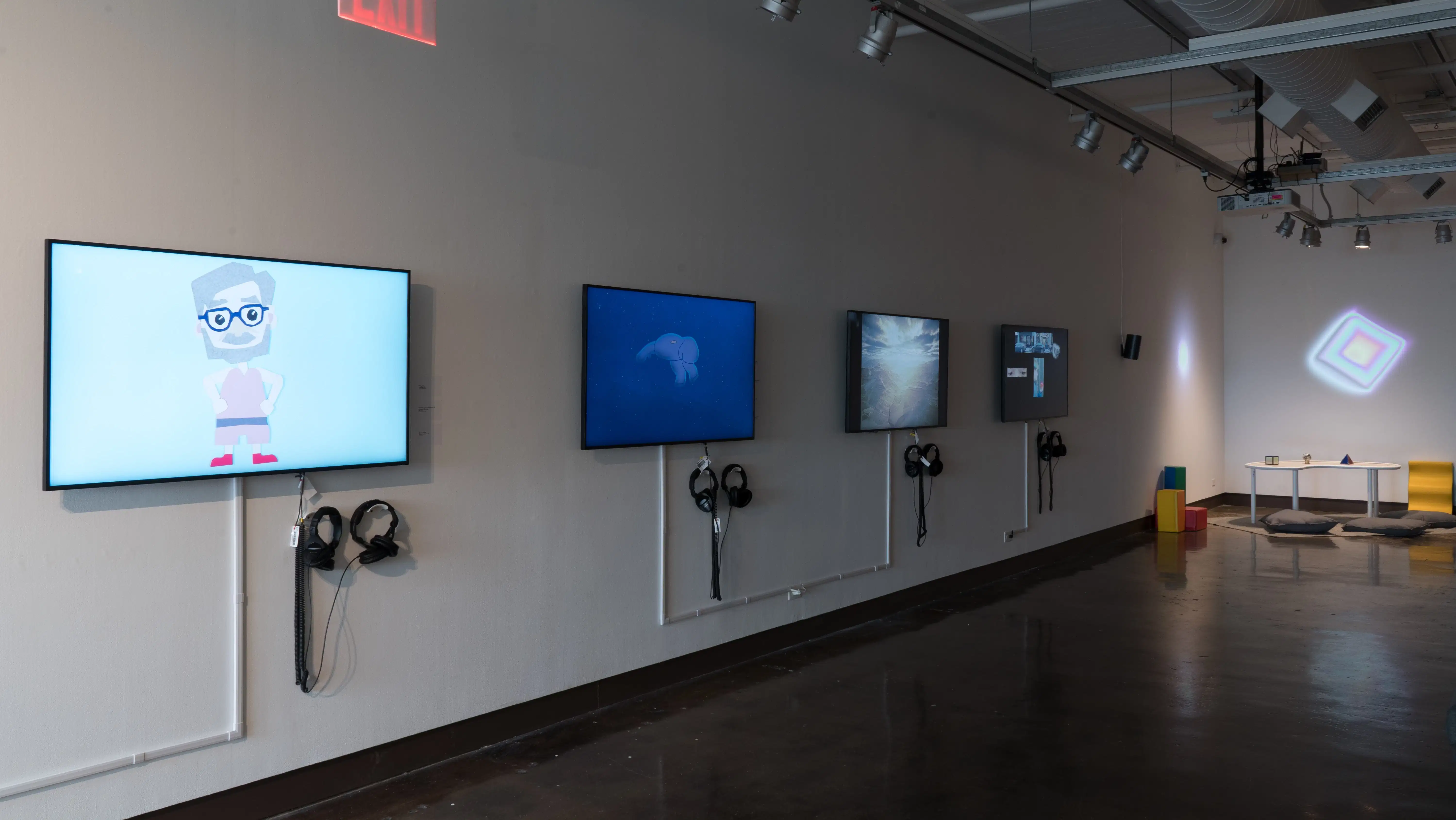 Four monitors along a wall in gallery one play many animated videos on a loop. In the corner to the right, there are colorful projections on the wall, and a small table with three differently shaped objects.