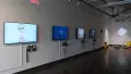 Four monitors along a wall in gallery one play many animated videos on a loop. In the corner to the right, there are colorful projections on the wall, and a small table with three differently shaped objects.