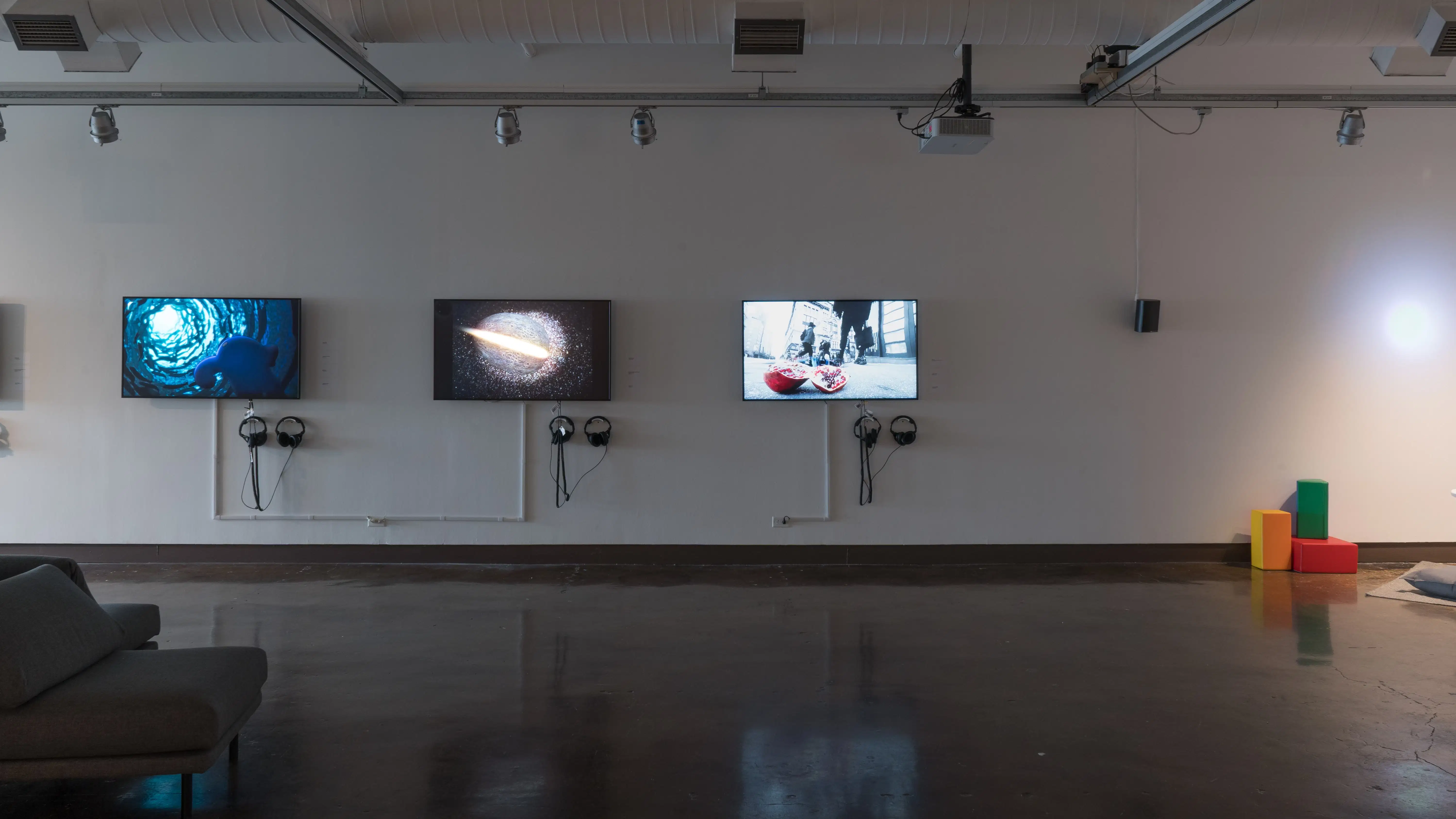 Three monitors along a wall in gallery one play many animated videos on a loop. Colorful blocks sit on the floor to the far right.