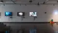 Three monitors along a wall in gallery one play many animated videos on a loop. Colorful blocks sit on the floor to the far right.