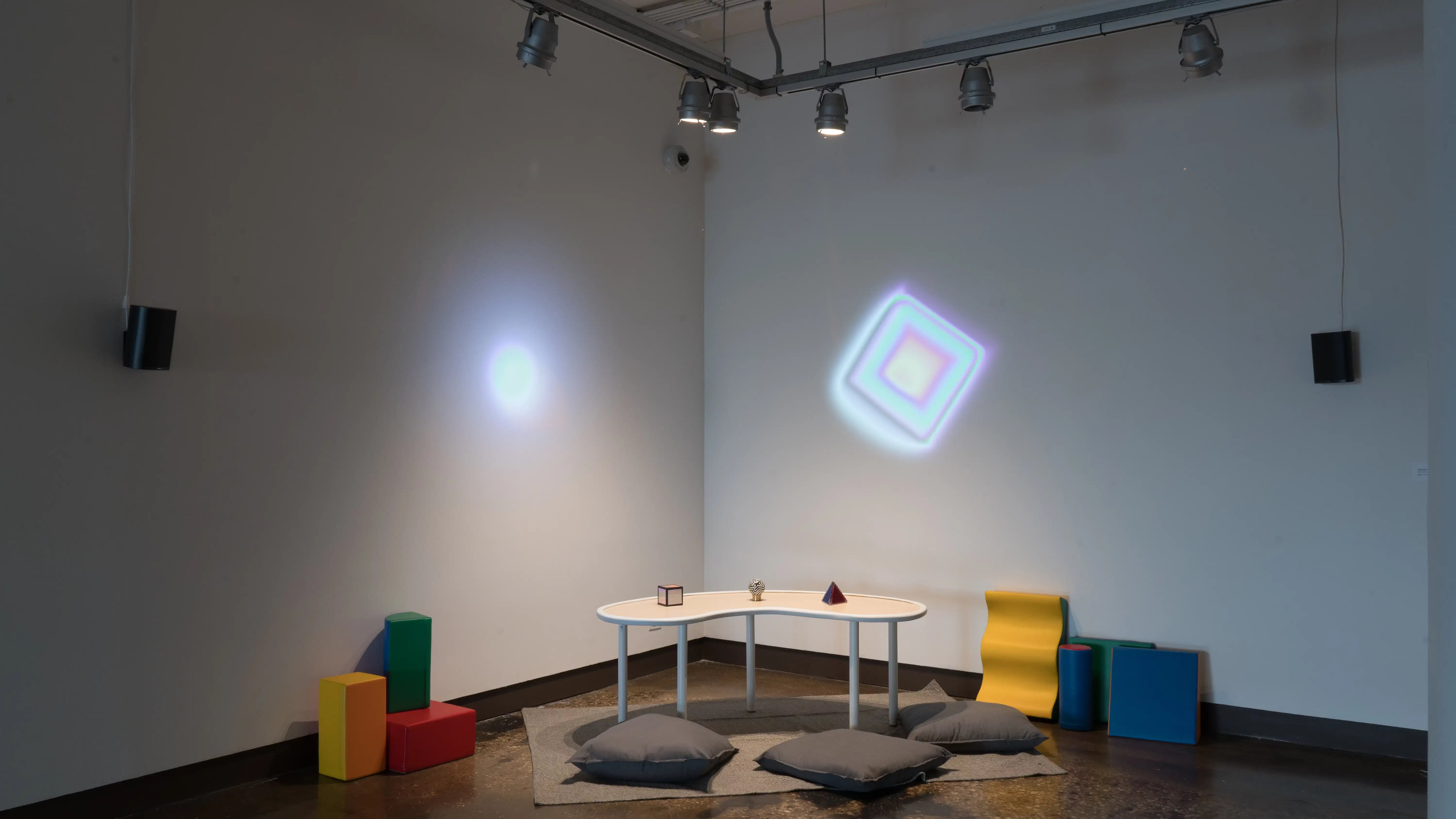 In a corner of gallery one, three objects (a cube, a sphere, and a pyramid) are laid out on a small table. The table is surrounded by floor pillows, and colorful projections glow on both walls.