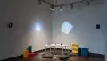 In a corner of gallery one, three objects (a cube, a sphere, and a pyramid) are laid out on a small table. The table is surrounded by floor pillows, and colorful projections glow on both walls.