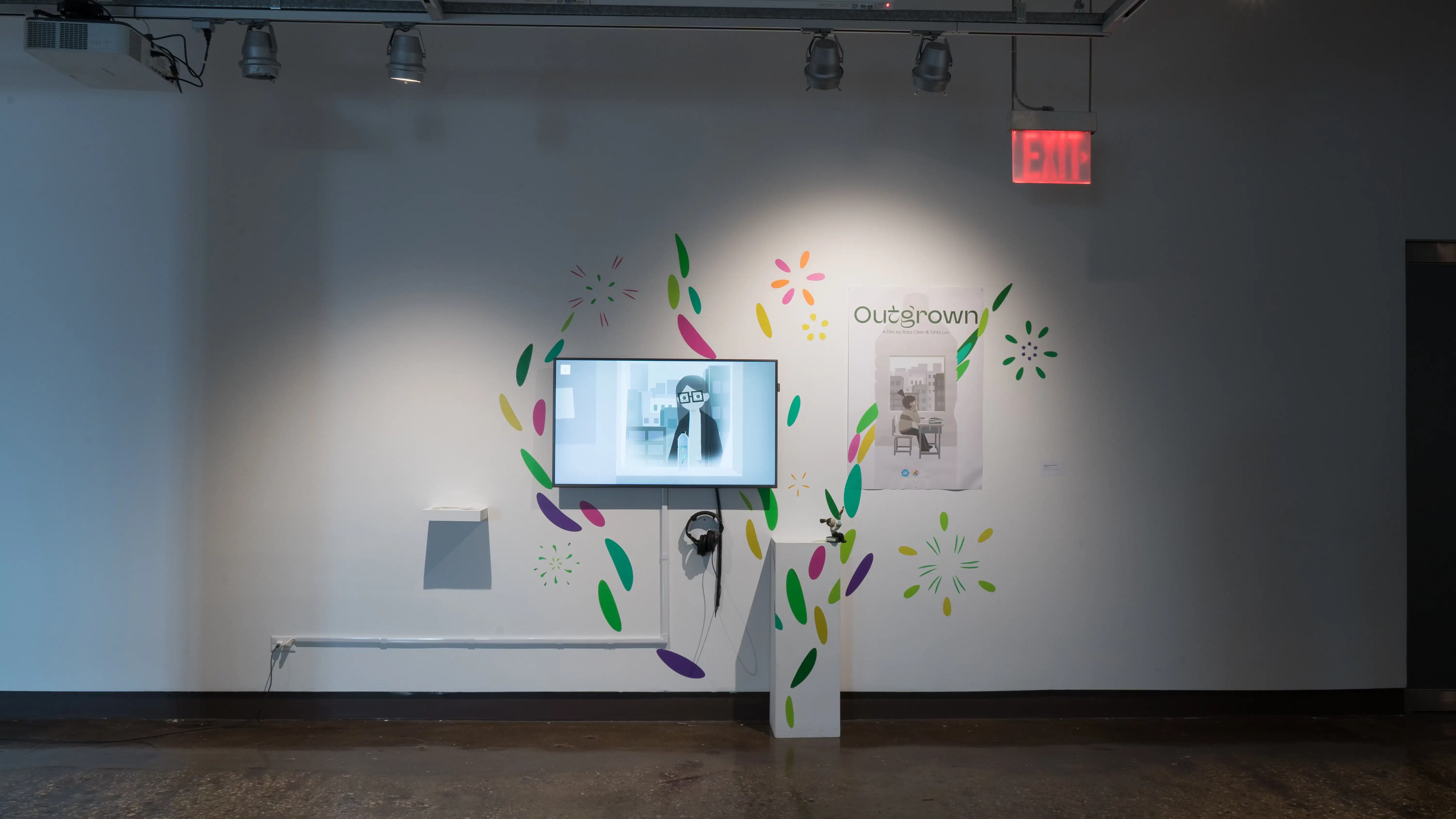 A monitor in gallery one is surrounded by colorful marks on the walls, which overflow onto the pedestal set in front of it. A poster beside the monitor reads “Outgrown.”