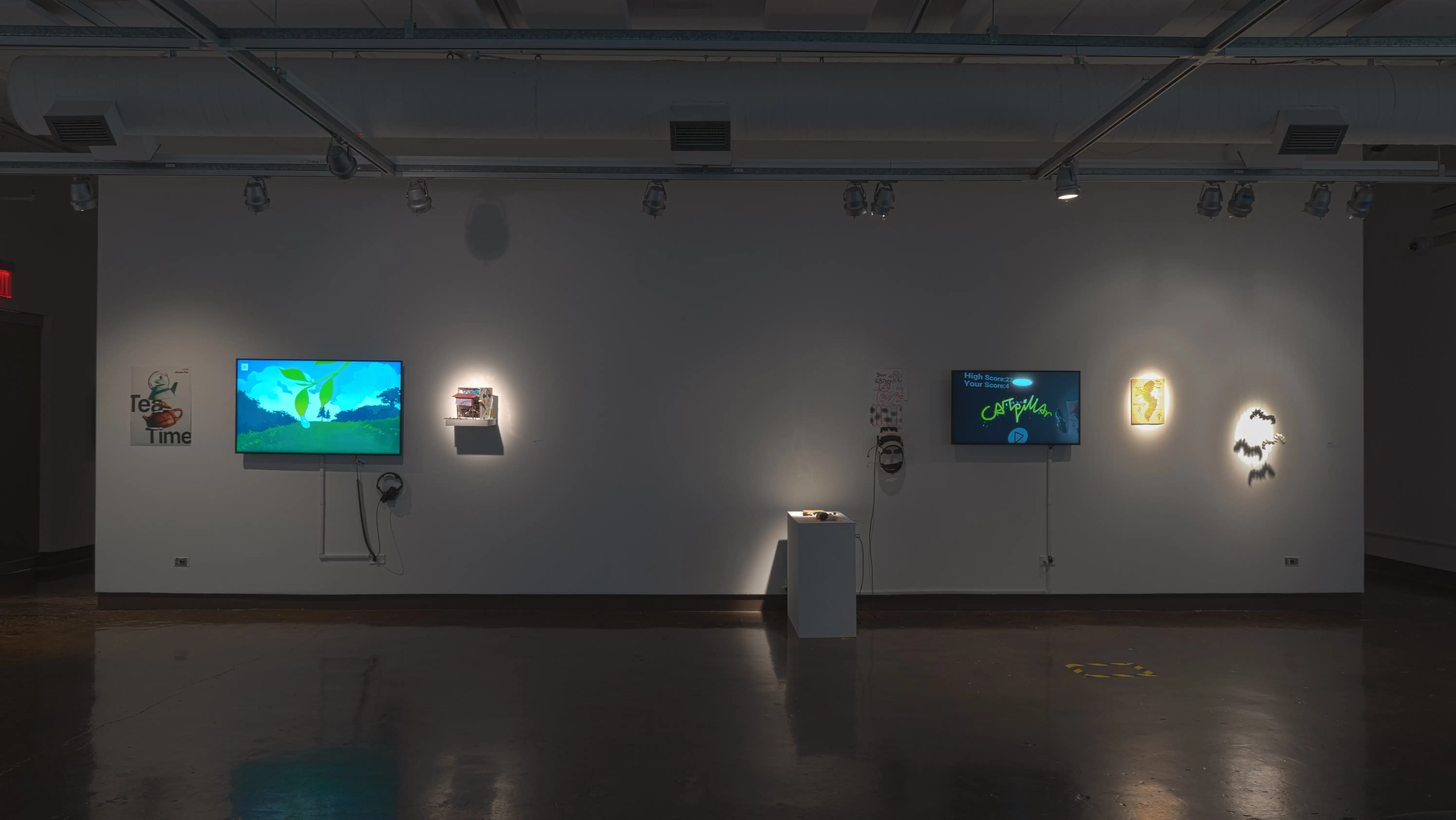 In gallery one, a monitor playing an animated video is flanked by a small poster and a shelf holding a small diorama. To the right, a VR video game is set up on a monitor, with a print and sculpture of caterpillars beside it.