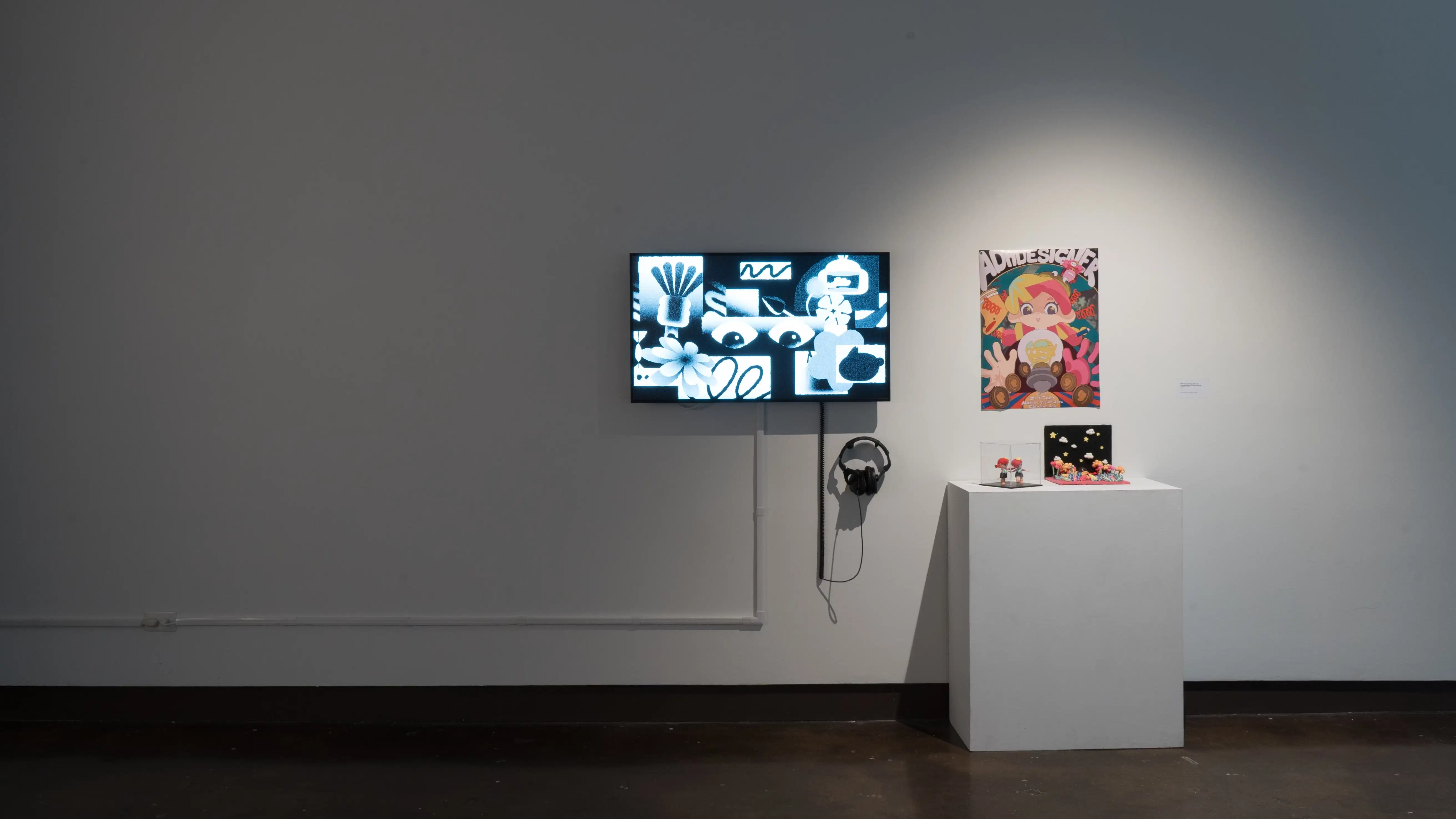 Beside a monitor playing an animated video, a white pedestal holds a small diorama and a tiny doll. A poster behind it depicts a girl with pink hair.