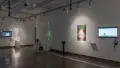 To the left of a TV monitor in gallery two is a colorful poster that reads “Super Metal Monkeys.” Beyond that is a projection featuring a green shape, and another monitor with a pedestal.