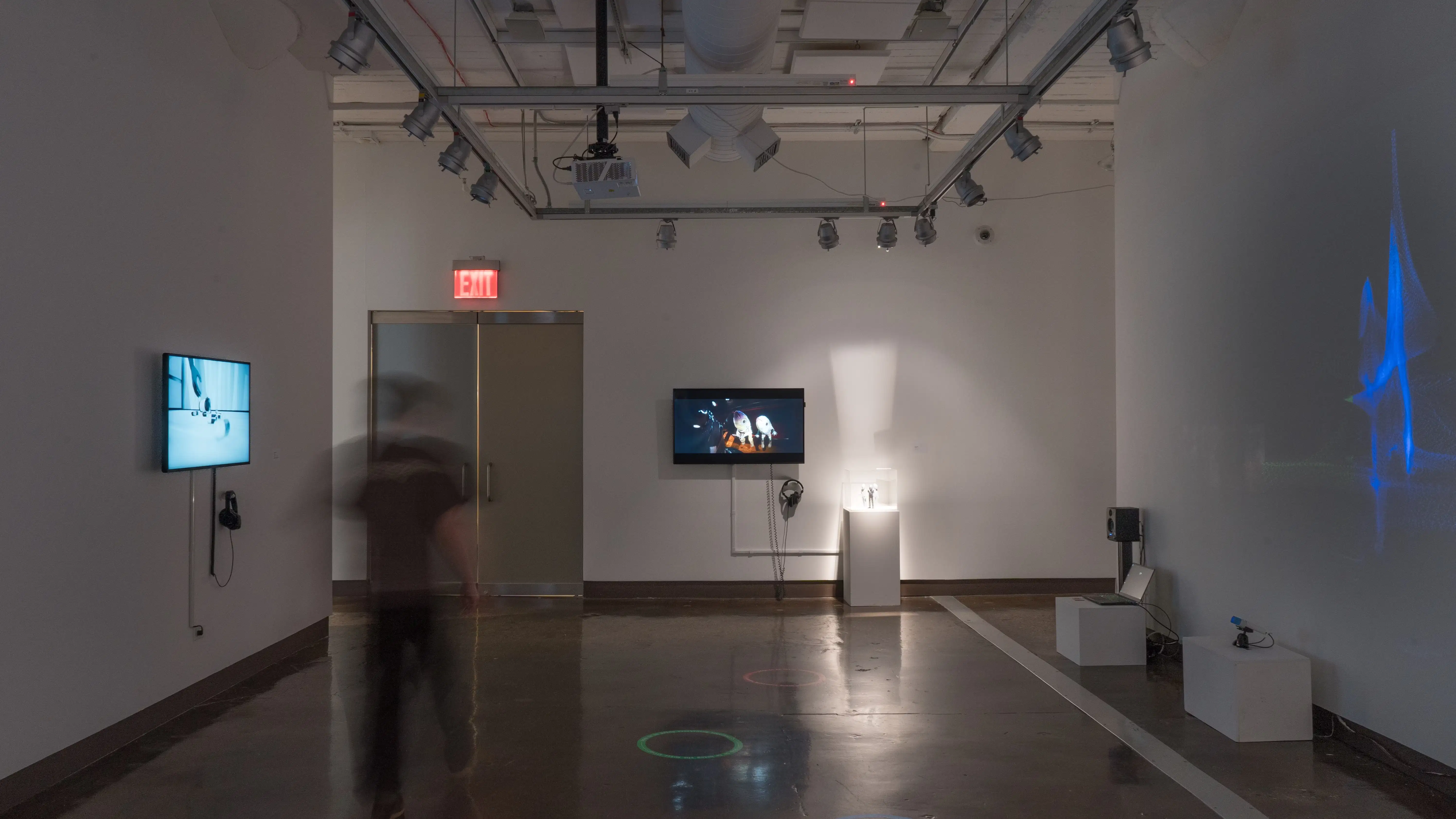 As a person walks through gallery two, a projection on the right wall reflects the movement with a blue shape. The left wall displays a TV monitor, while the wall straight ahead displays a monitor and a pedestal.