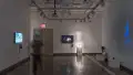 As a person walks through gallery two, a projection on the right wall reflects the movement with a blue shape. The left wall displays a TV monitor, while the wall straight ahead displays a monitor and a pedestal.