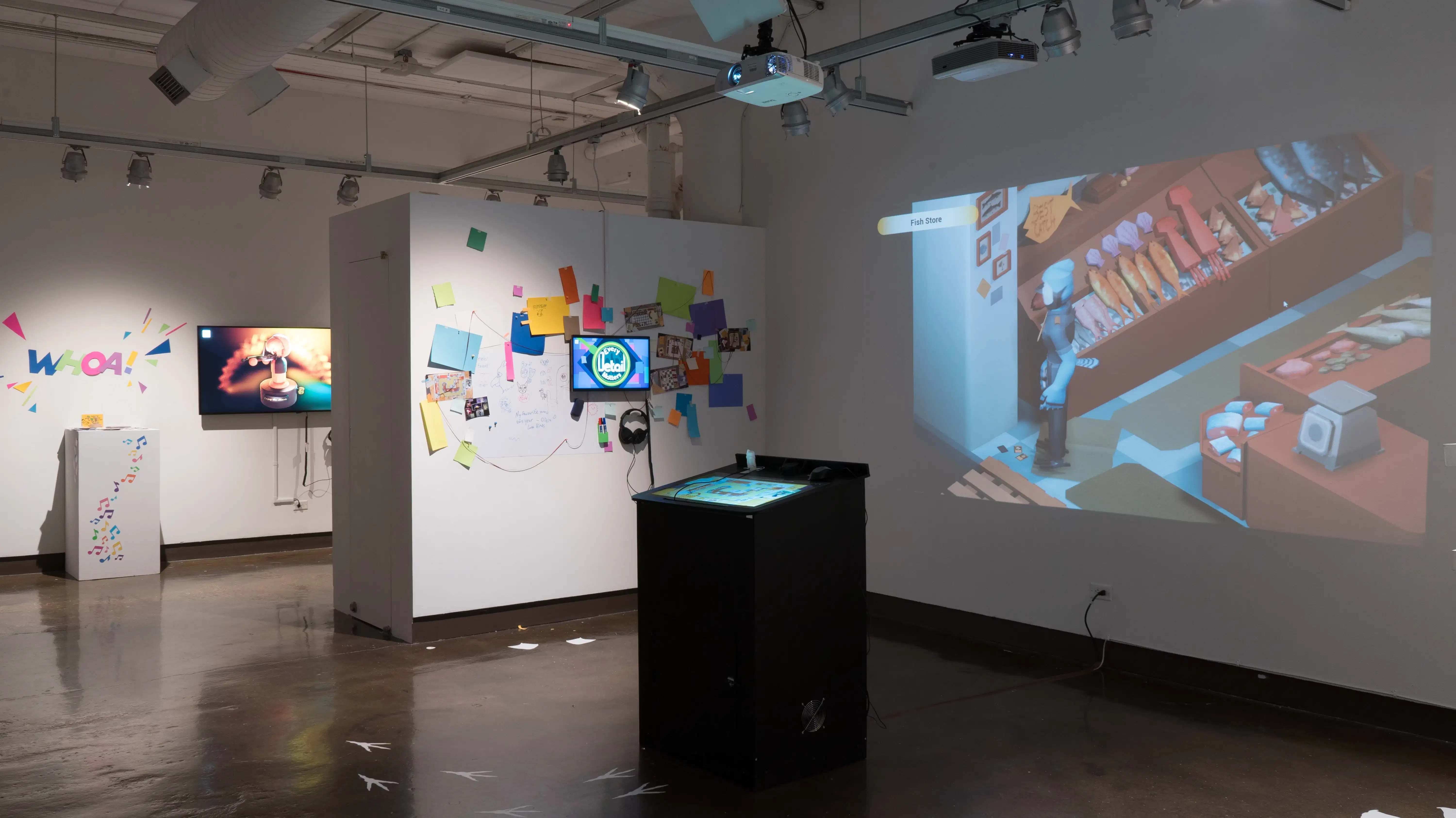 A small kiosk sits in the center of the gallery three alcove as a video game is projected on the wall behind it. On the side wall, a TV monitor plays an animated video, and many pieces of colorful paper surround the monitor.