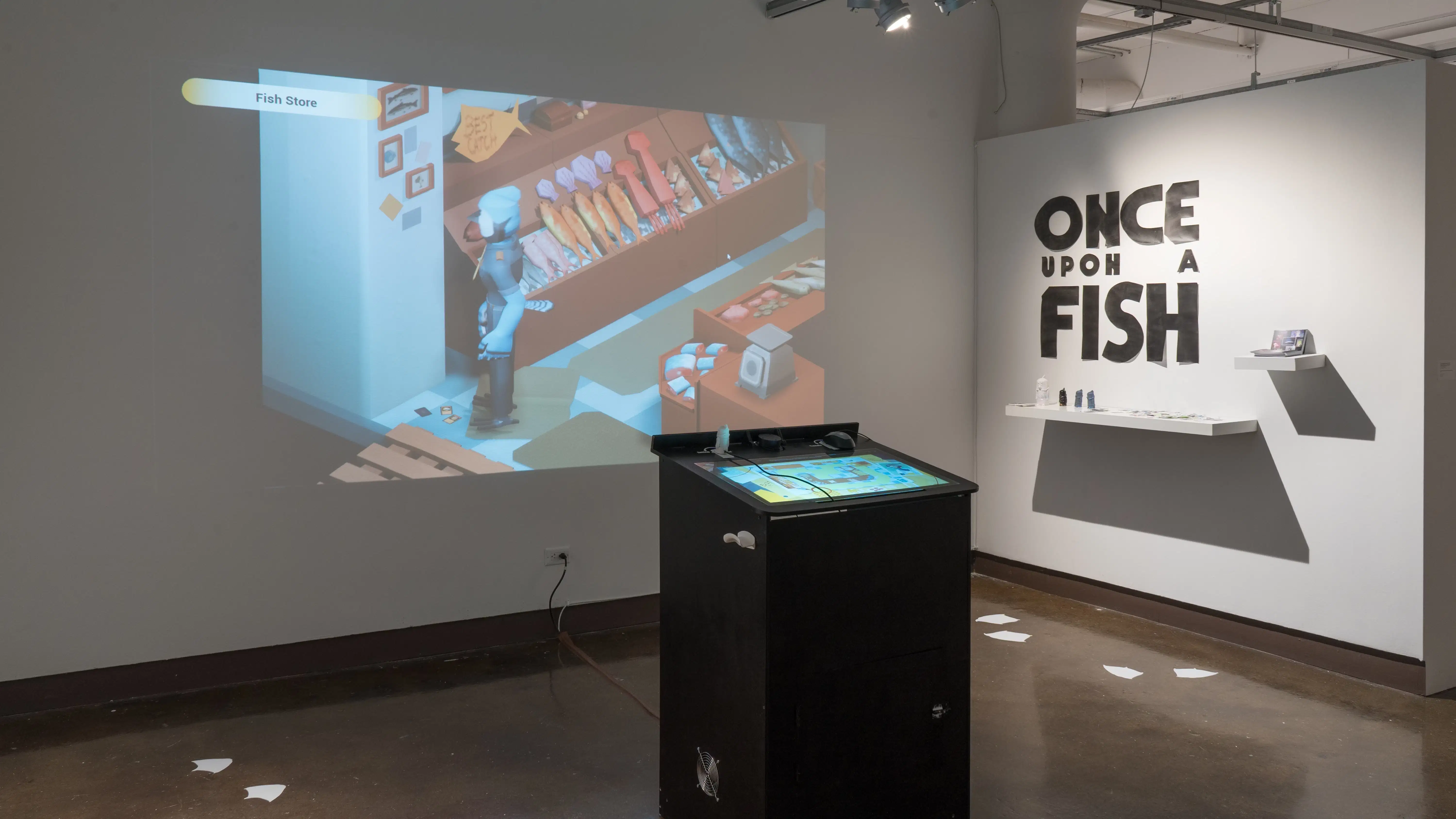 In the gallery three alcove, bold text on one wall reads “Once Upon A Fish.” A black kiosk in the center of the space has a screen to play a video game. The game is projected onto the wall behind the kiosk.