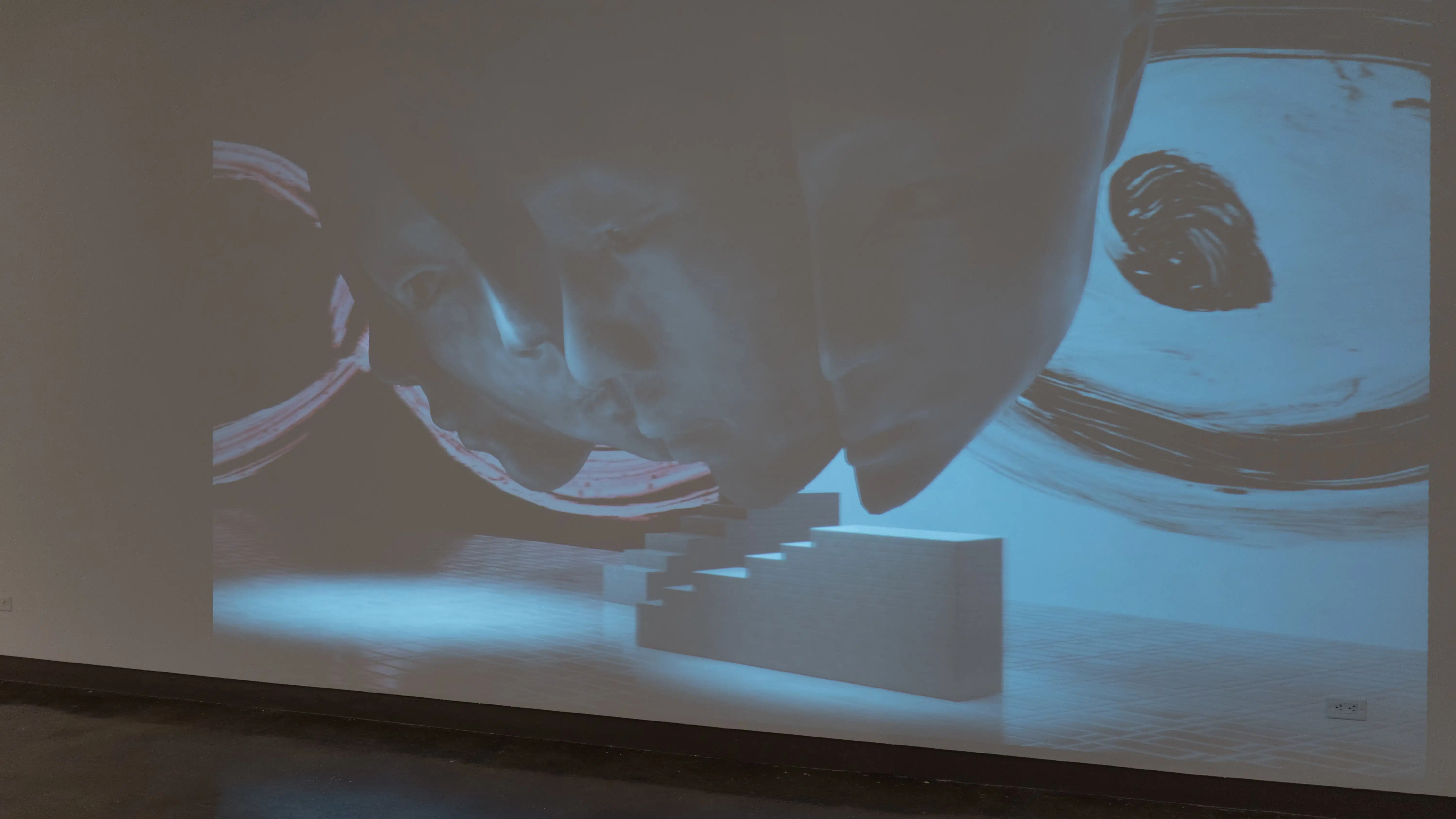 A blue and black projection depicting a mask and some stairs is shown on the wall of gallery three.