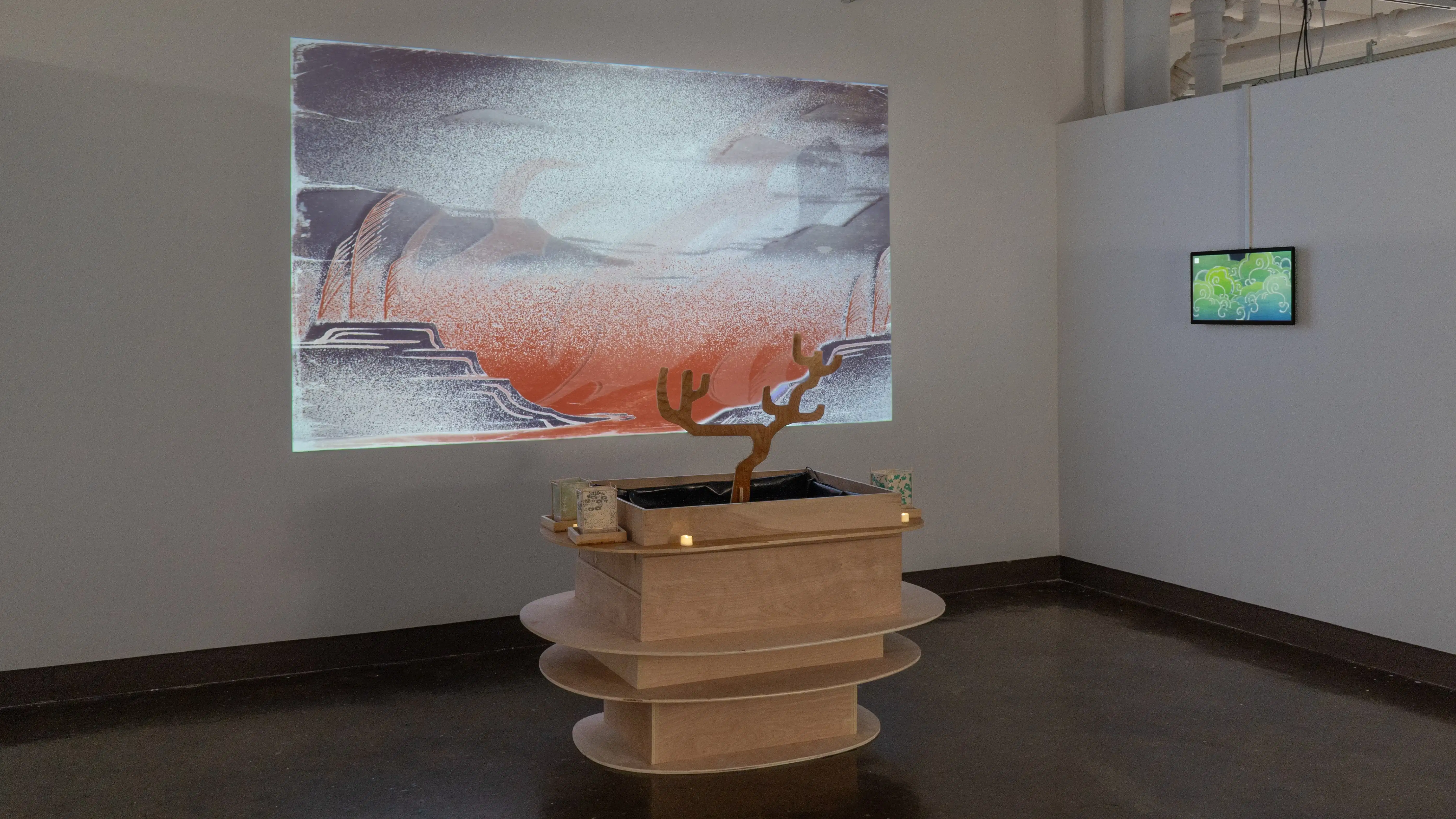 A wooden sculpture of square boxes tiered with oval-shaped shelves sits on the floor in the center of the gallery four alcove. Small lanterns and votive candles sit on the shelf around the top of the sculpture, and an illustrated landscape is projected on the back wall.