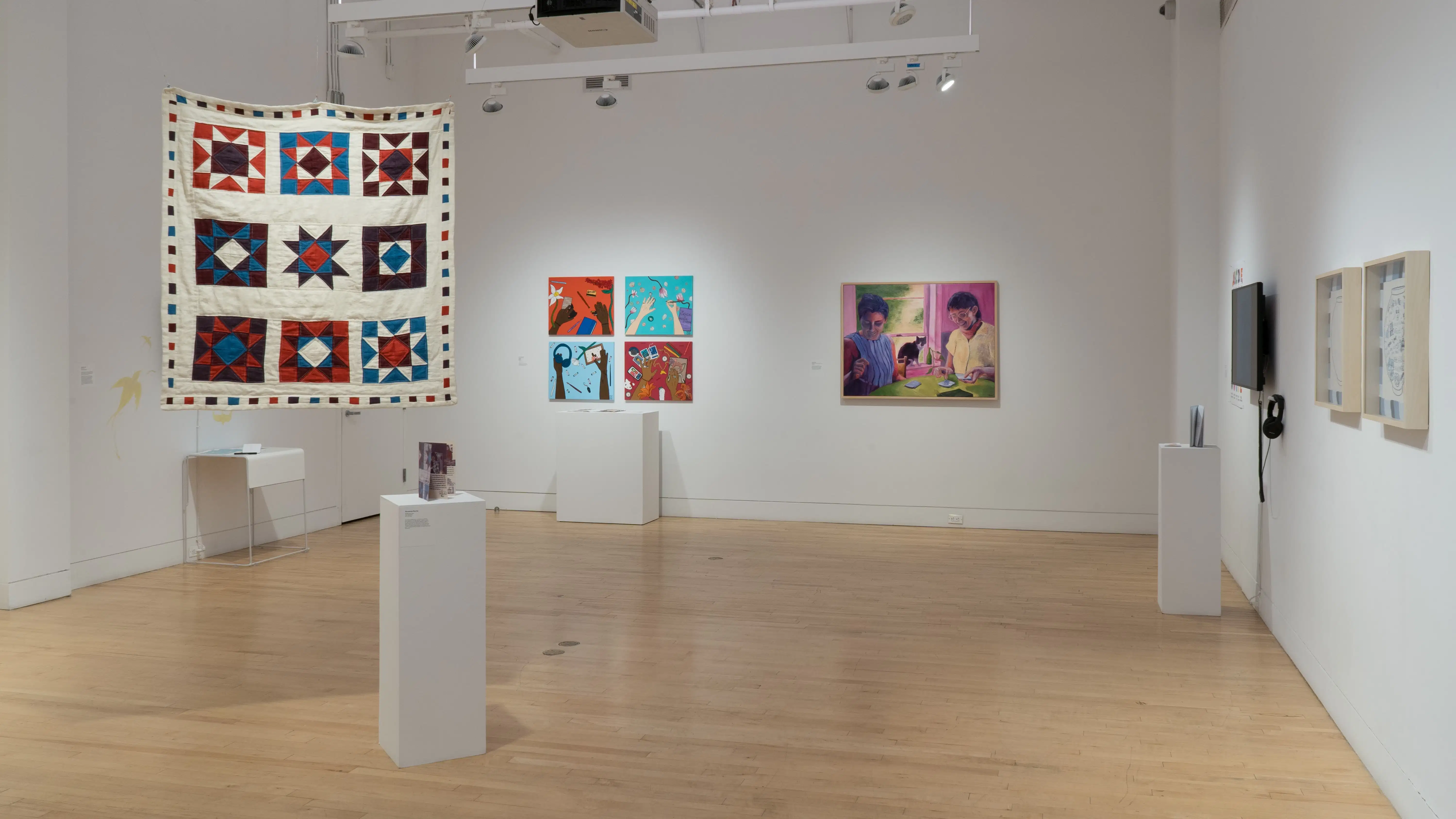 Overview shot of the SVA Flatiron Gallery, showing a large square quilt hanging near a pedestal in the foreground, with a grid of paintings and a large painting on the back wall next to each other. 