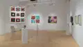 Overview shot of the SVA Flatiron Gallery, showing a large square quilt hanging near a pedestal in the foreground, with a grid of paintings and a large painting on the back wall next to each other. 