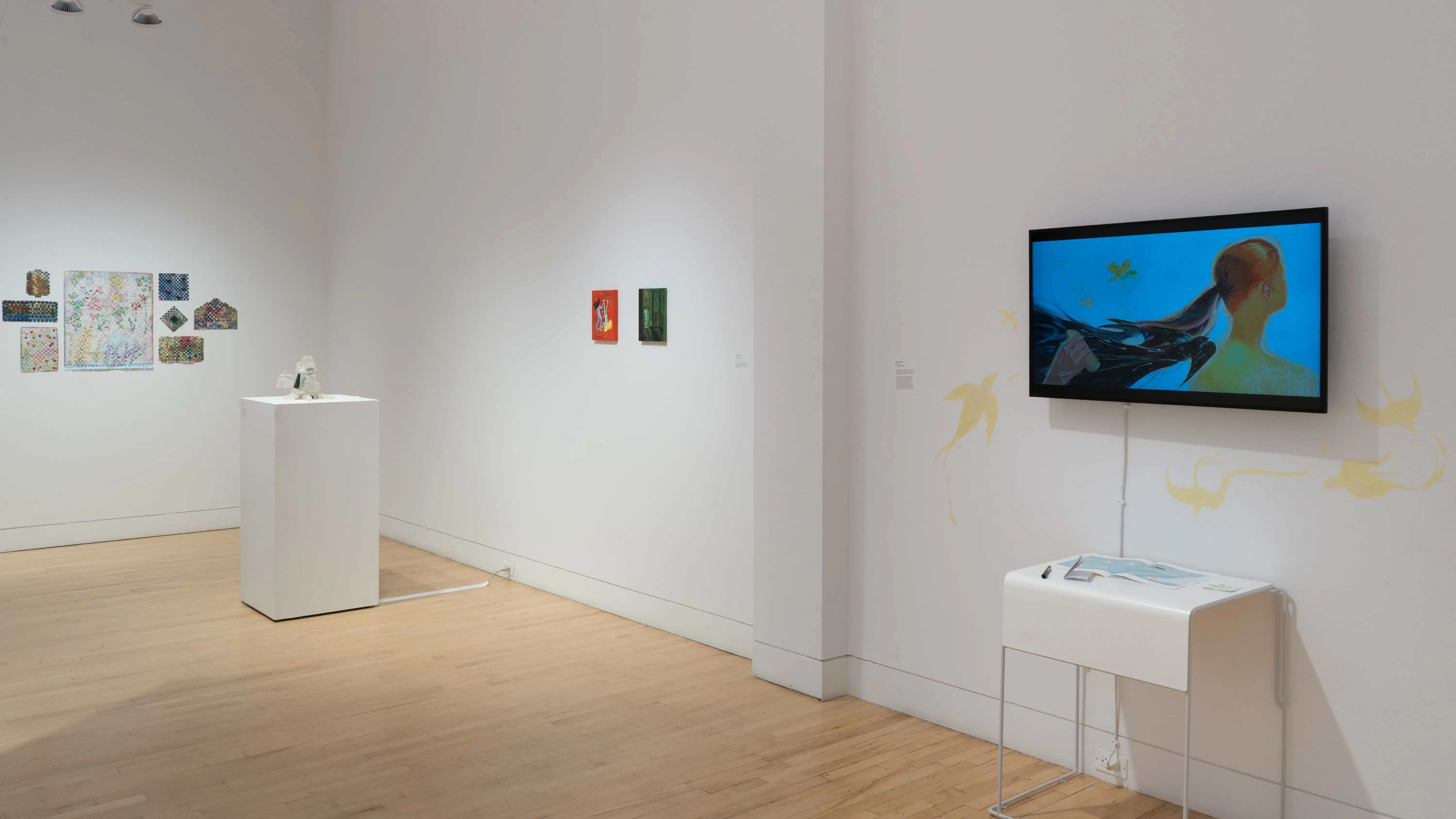 Angled shot of the west wall at the SVA Flatiron Gallery, showing a monitor with a figure’s hair blowing in the wind in the foreground. There is a pedestal with a small sculpture in the background.