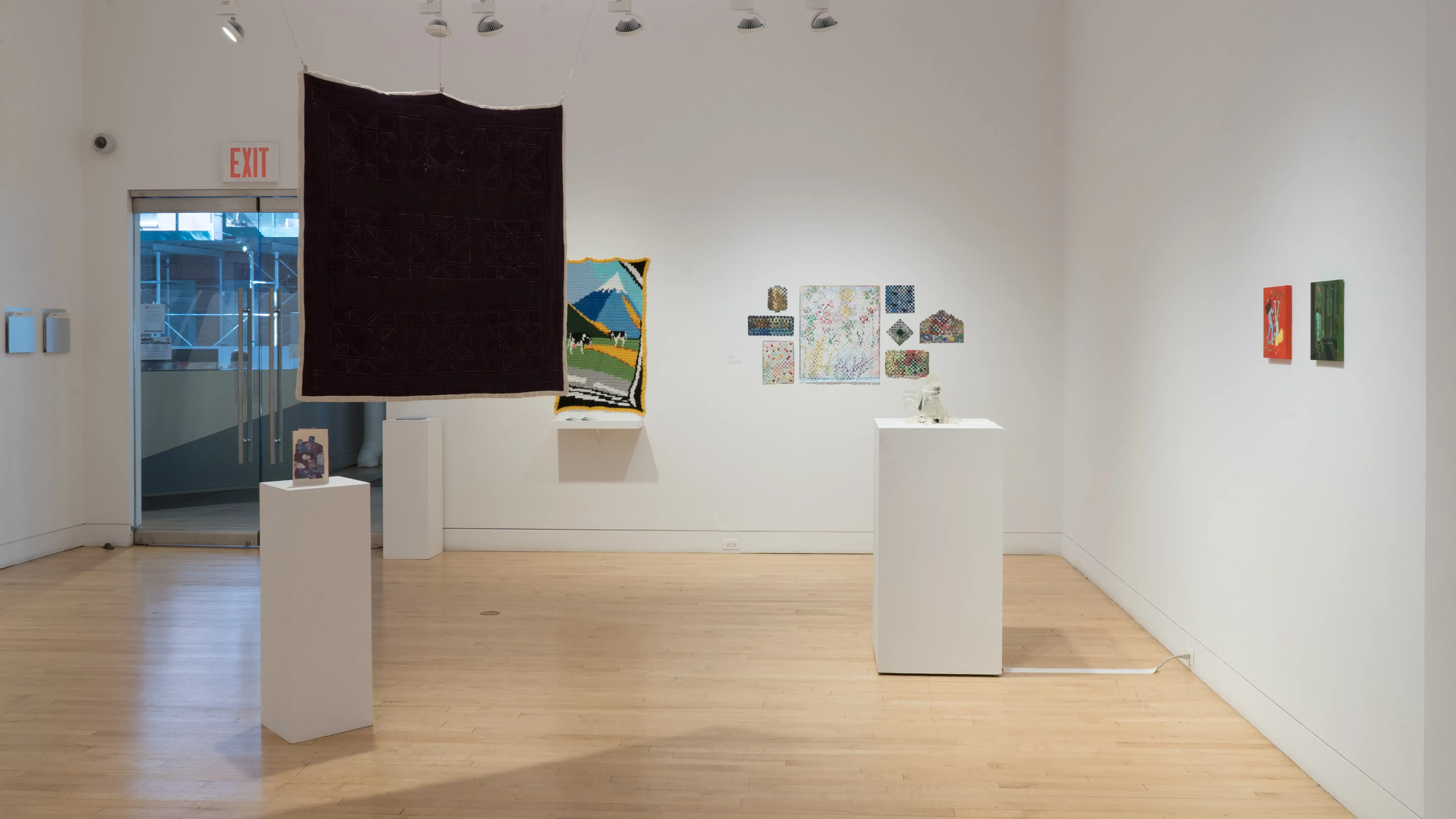 Straight shot of the south wall at the SVA Flatiron Gallery, showing a large square quilt hanging near a pedestal in the foreground and a pedestal with a small sculpture in the background.