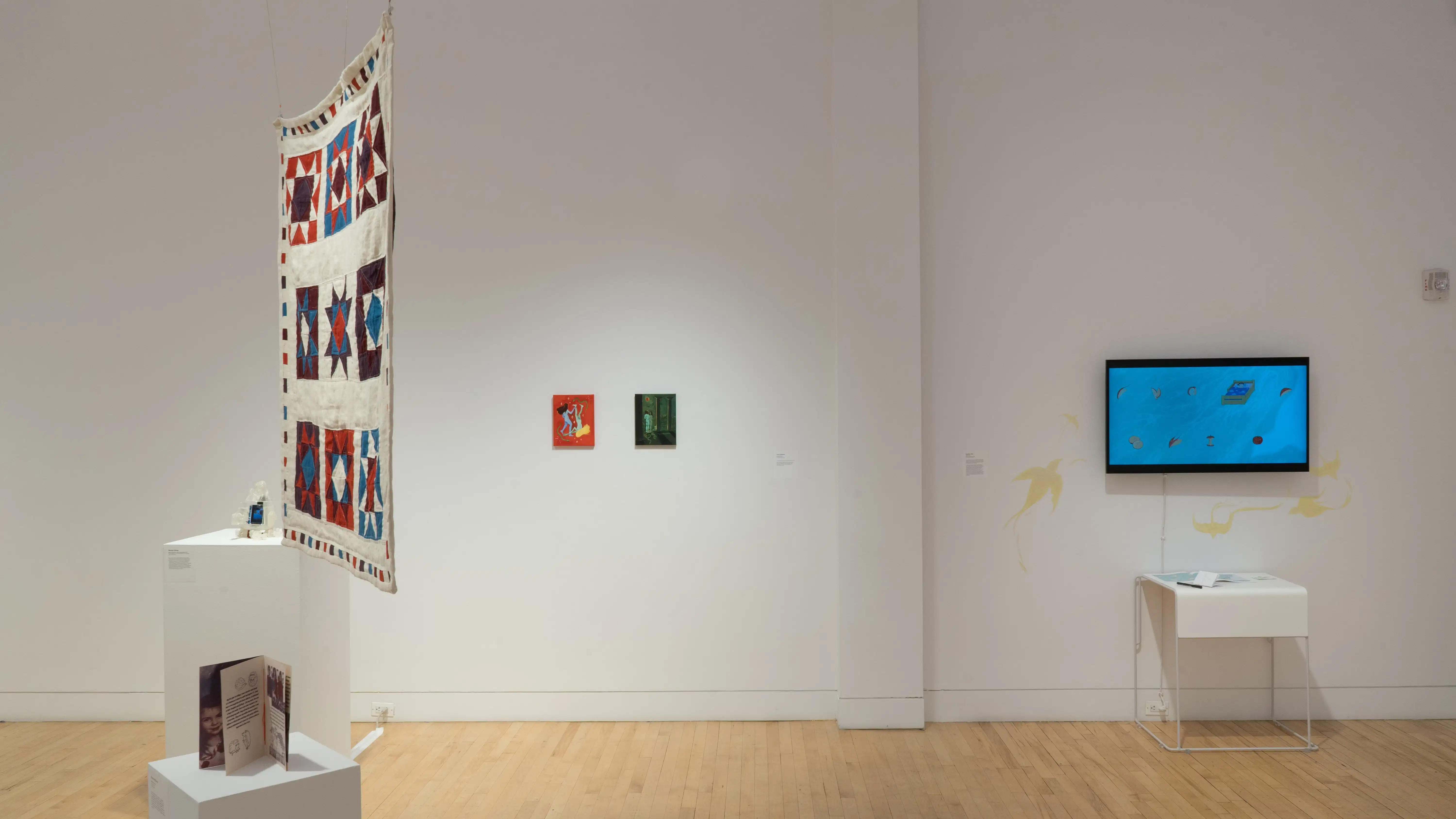 Straight shot of the west wall at the SVA Flatiron Gallery, showing a monitor with a figure’s hair blowing in the wind on the right, a large square quilt hanging near a pedestal in the foreground, and two small paintings in the background.