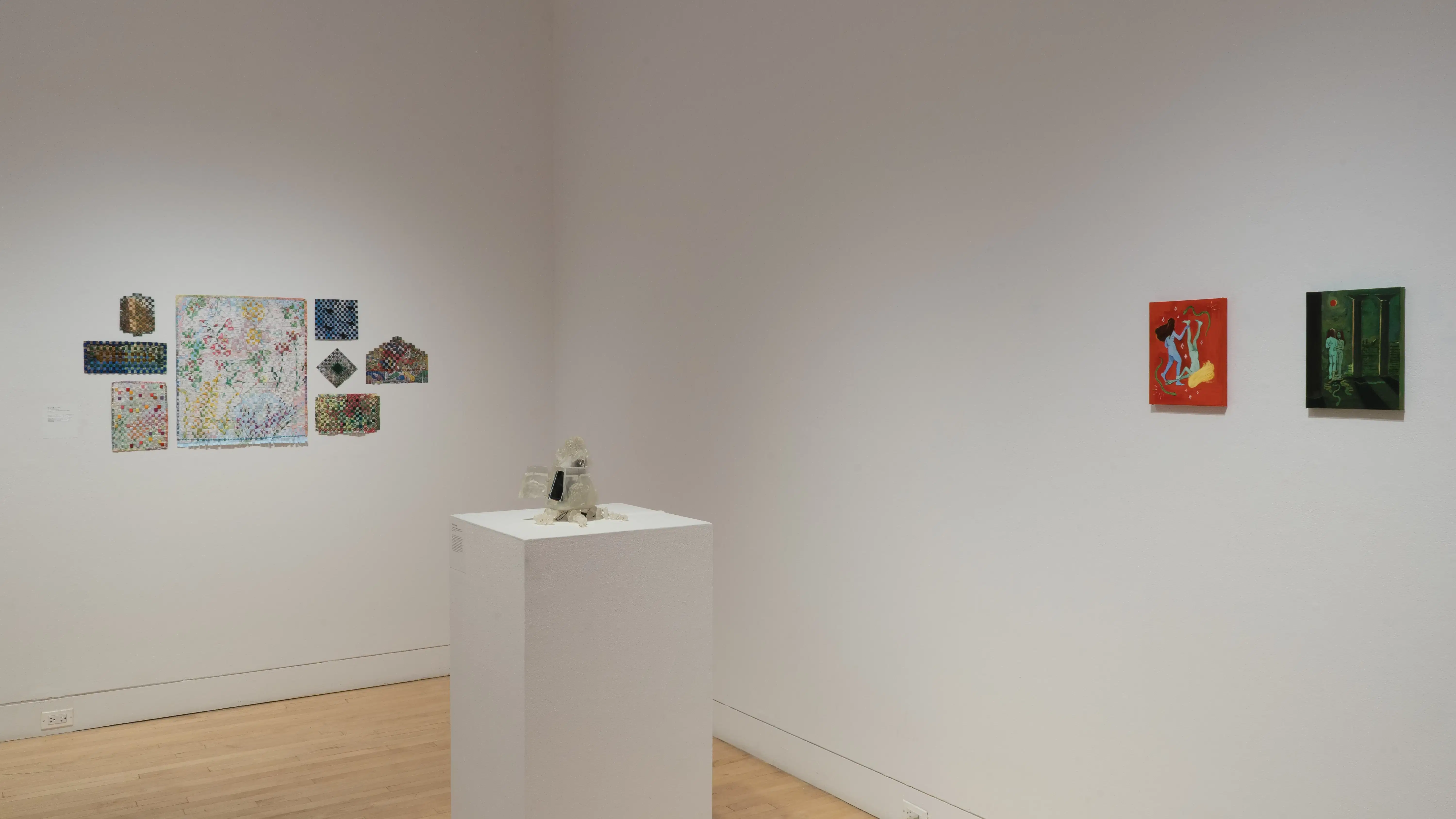 Angled view of the southwest corner of the SVA Flatiron Gallery, showing a pedestal with a small sculpture in the foreground, two small paintings on the wall in the back right, and a salon-style hanging of quilted paper works on the left. 