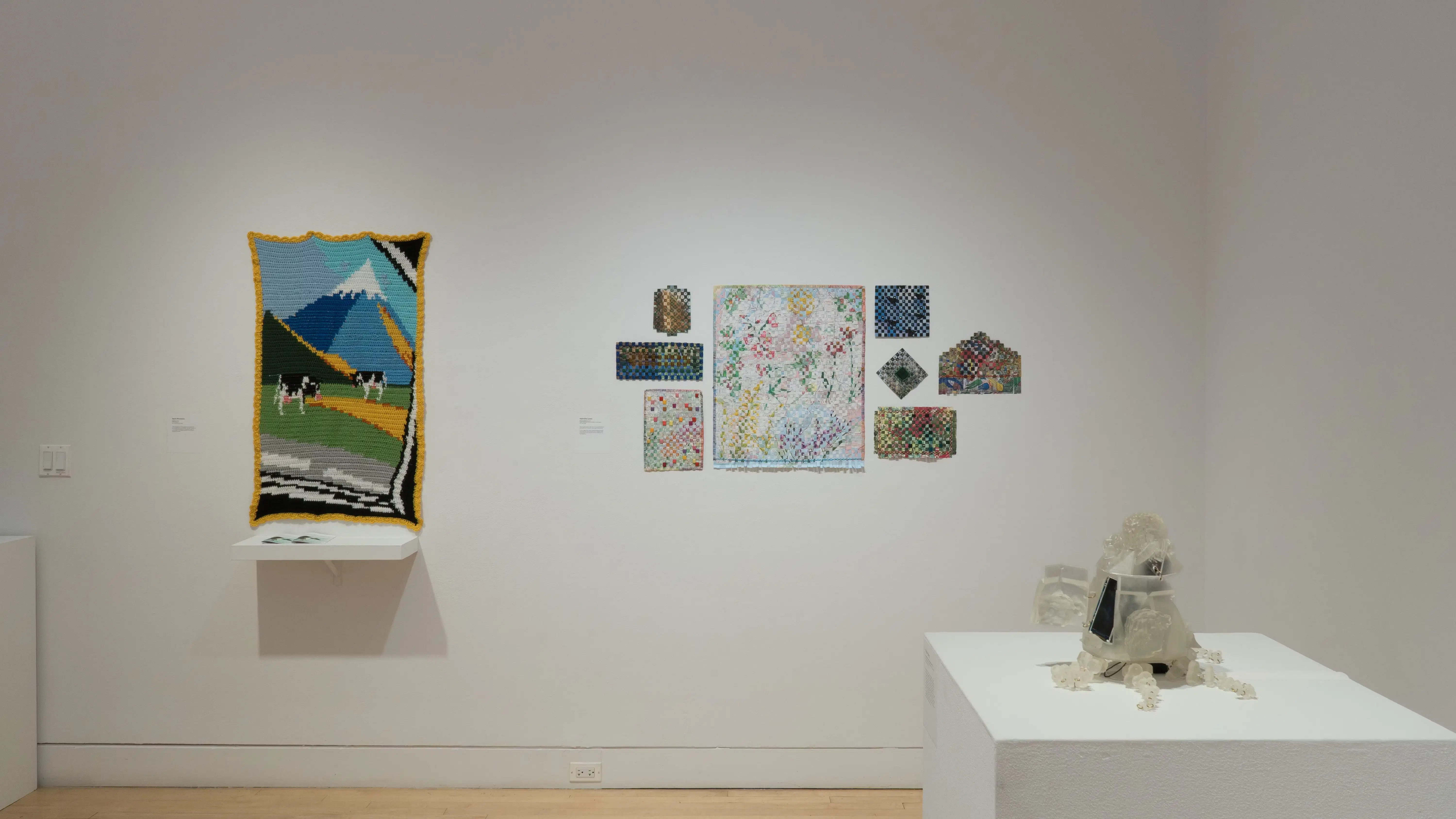 Straight view of the southwest corner of the SVA Flatiron Gallery, showing a pedestal with a small sculpture in the foreground, a crocheted mountain scene on the left, and a salon-style hanging of quilted paper works on the right. 
