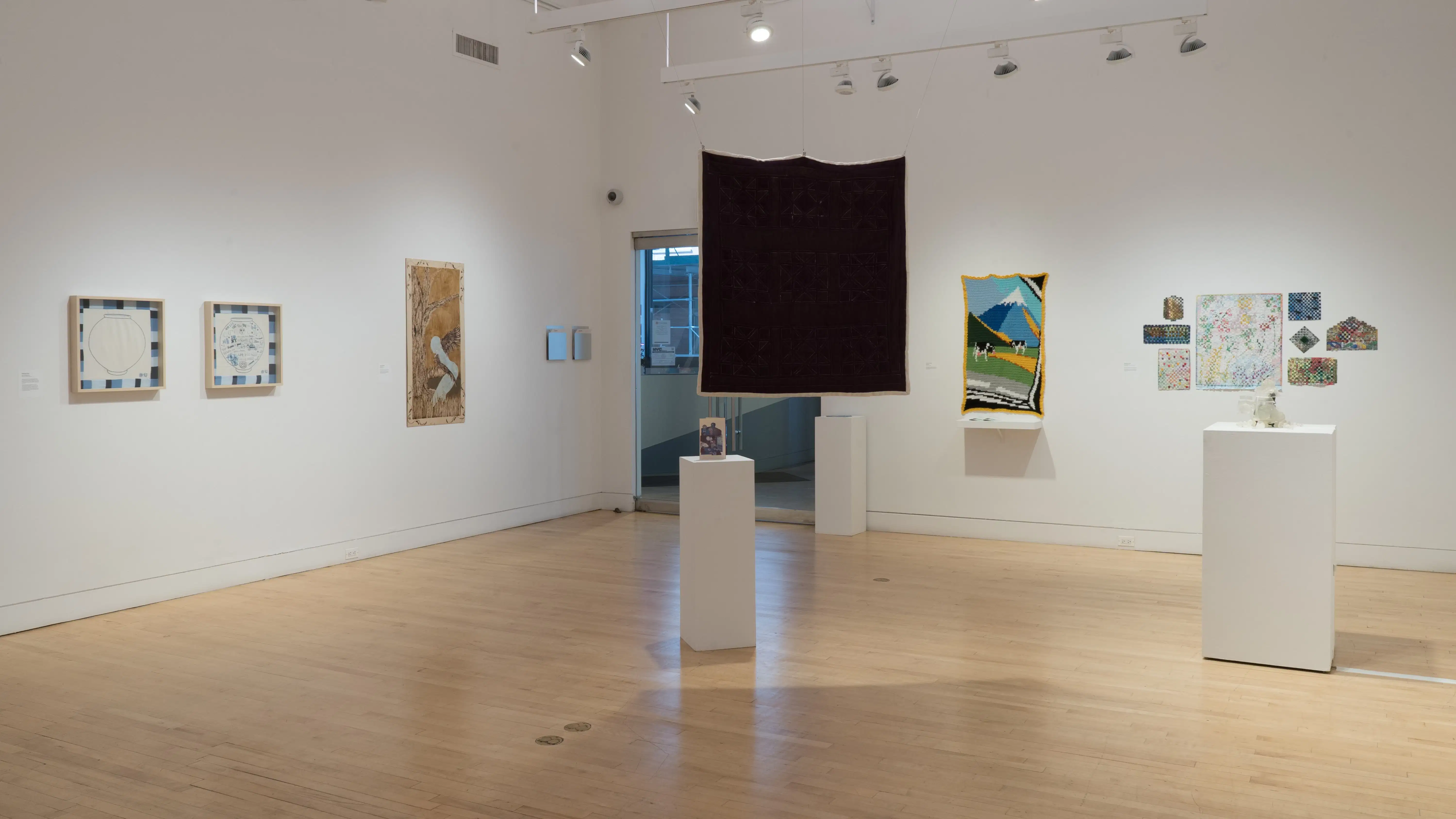 Overview shot of the SVA Flatiron Gallery from the back, showing a large square quilt hanging near a pedestal in the foreground, with a grid of paper works on the back wall, a crocheted mountain scene on the left, and some framed works on the east wall.