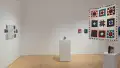 Straight view of the southwest corner of the SVA Flatiron Gallery, showing a pedestal with a small sculpture behind a square quilt in the foreground, and salon-style hanging of quilted paper works on the left. 