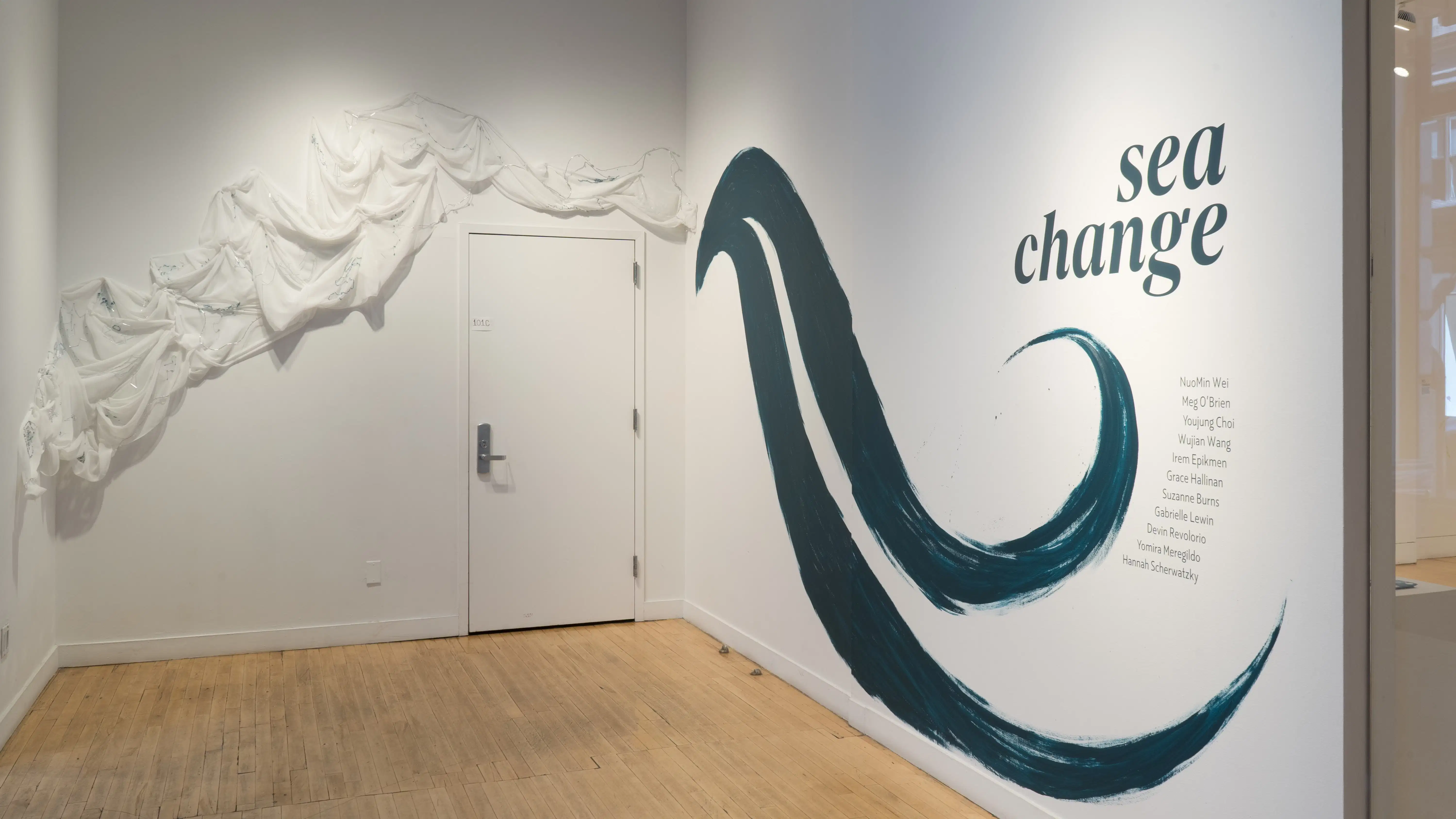Angled view of the alcove at the SVA Flatiron Gallery, showing a painted wall with a wave and the words “sea change” to the right and a textile work along the wall and over the door on the left.