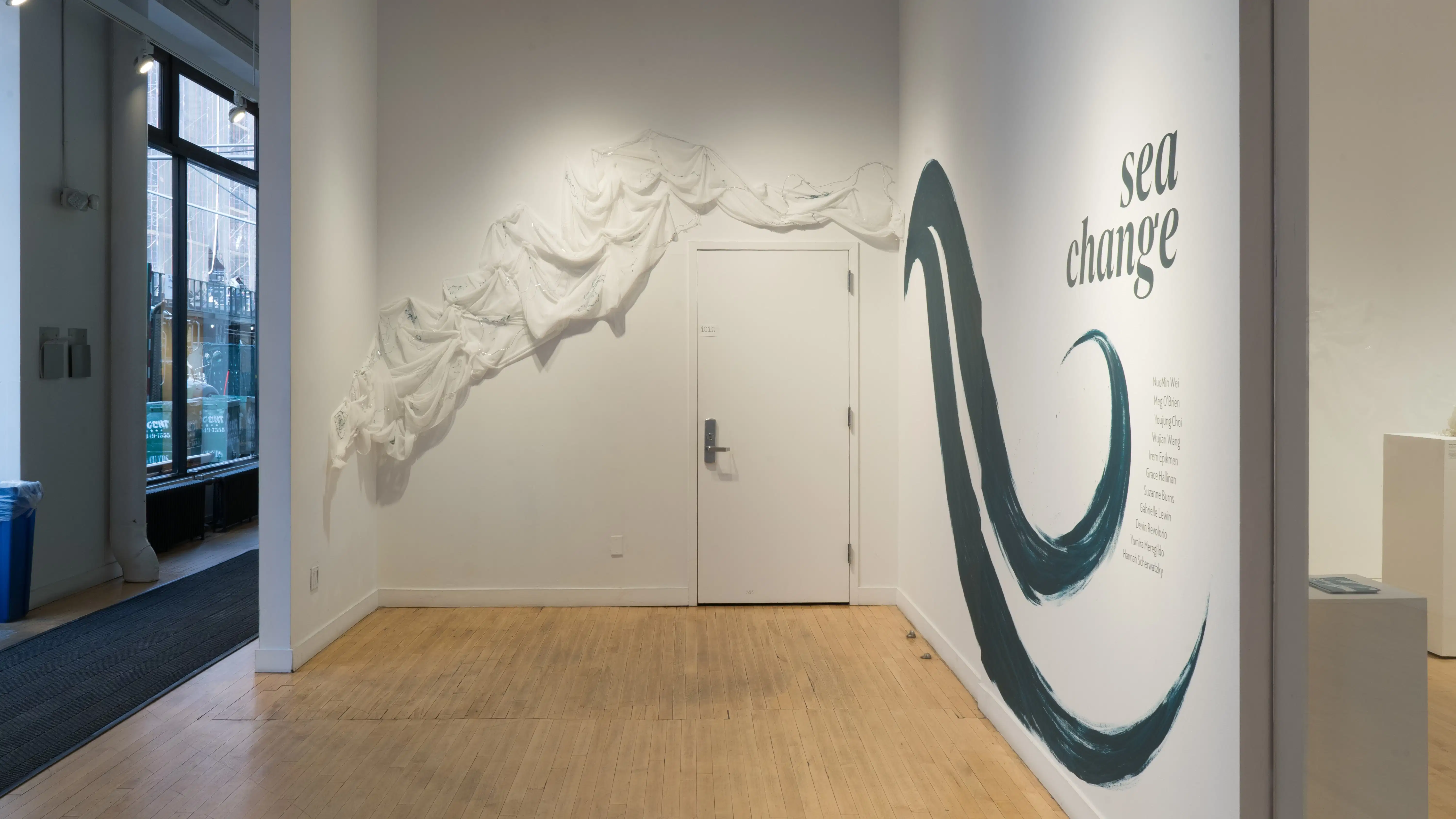 Straight view of the alcove at the SVA Flatiron Gallery, showing a painted wall with a wave and the words “sea change” to the right and a textile work along the wall and over the door on the left.