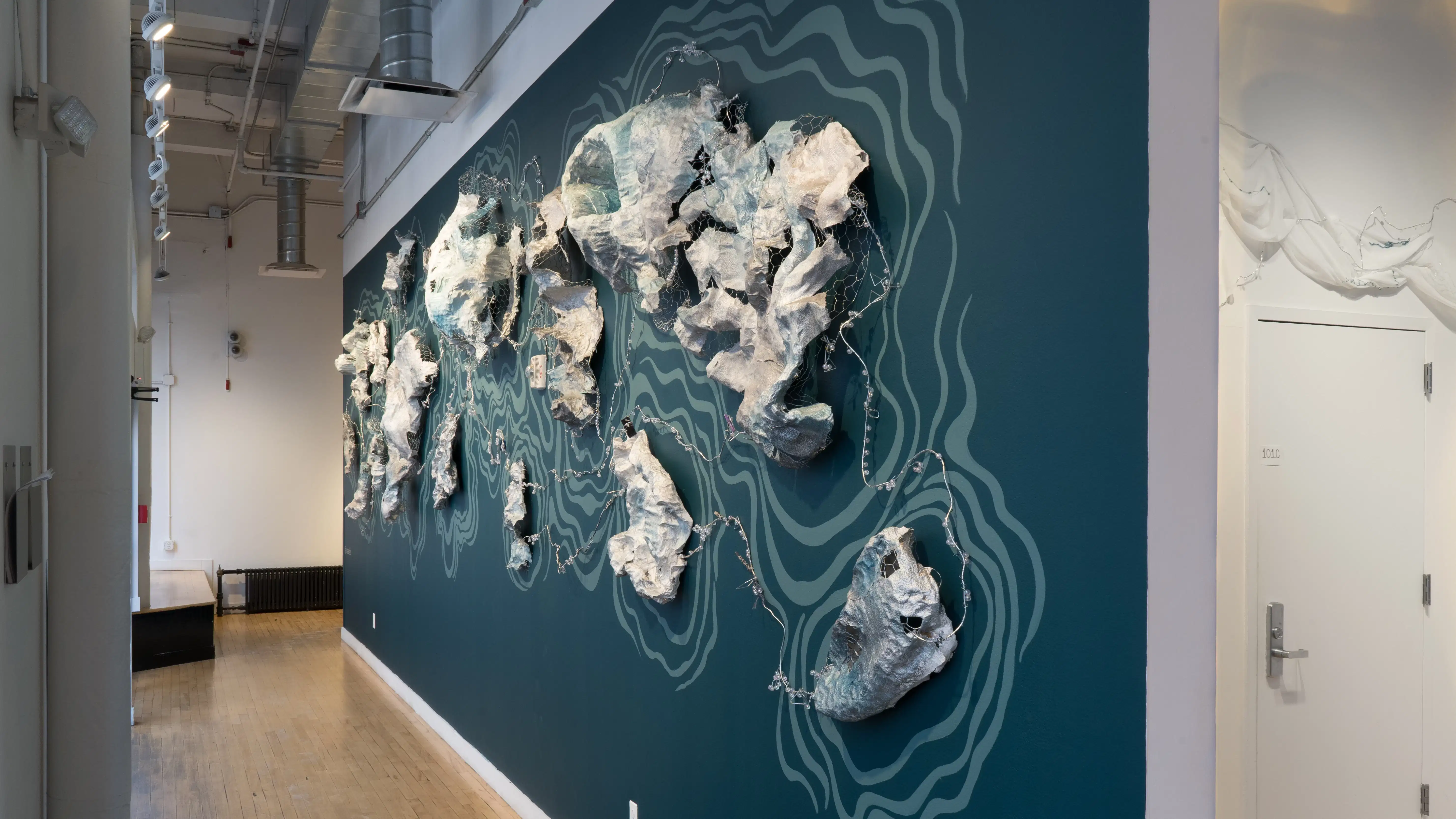 Angled view of the window wall at the SVA Flatiron Gallery facing west, showing a sculptural wall installation resembling sea creatures against a blue backdrop. 