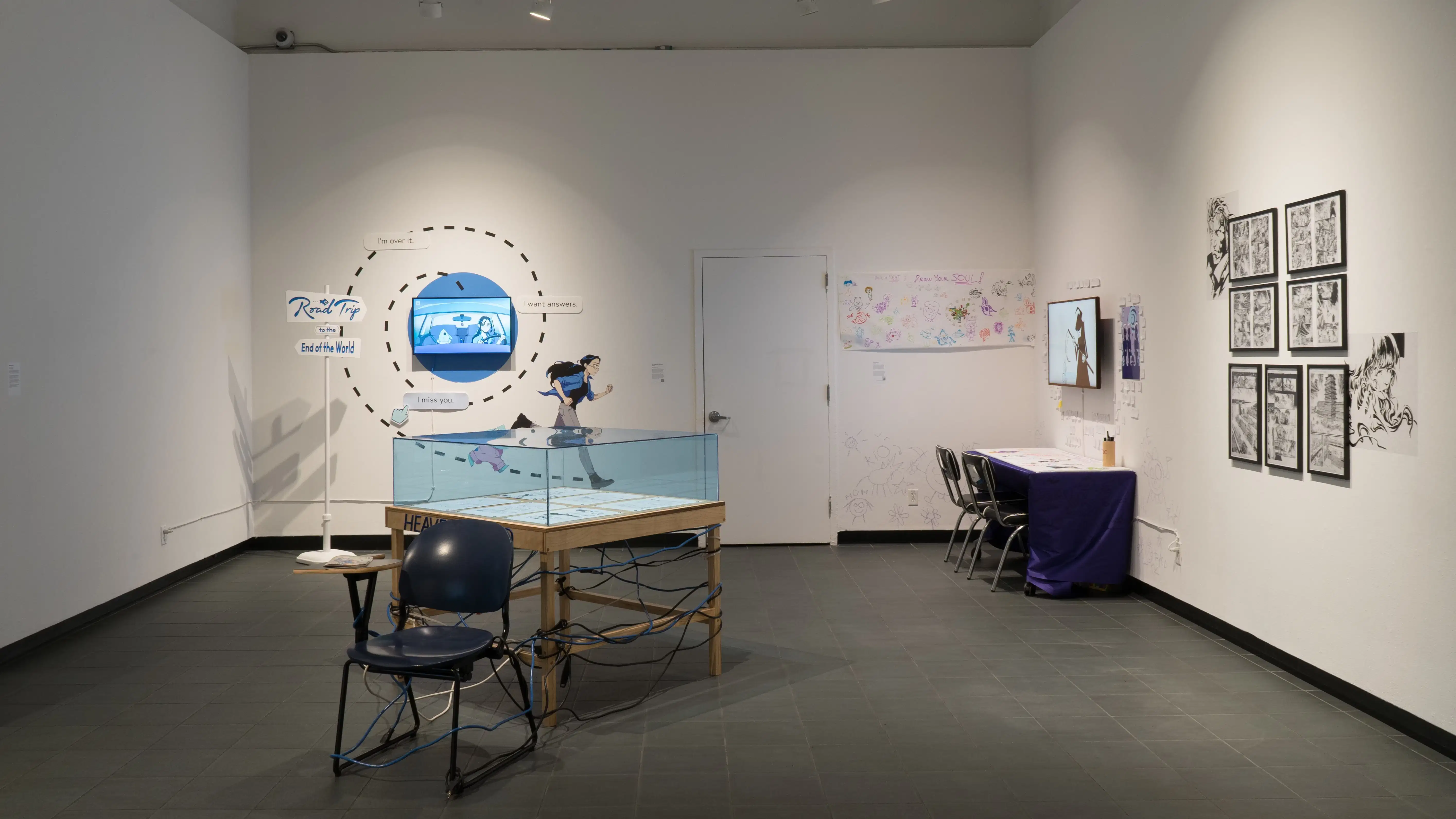 Several works in the ‘Now and Then exhibition. In the front is a glass display on a wooden table, with artworks present inside. Behind that are two animations by students and frames featuring black and white comic panels.