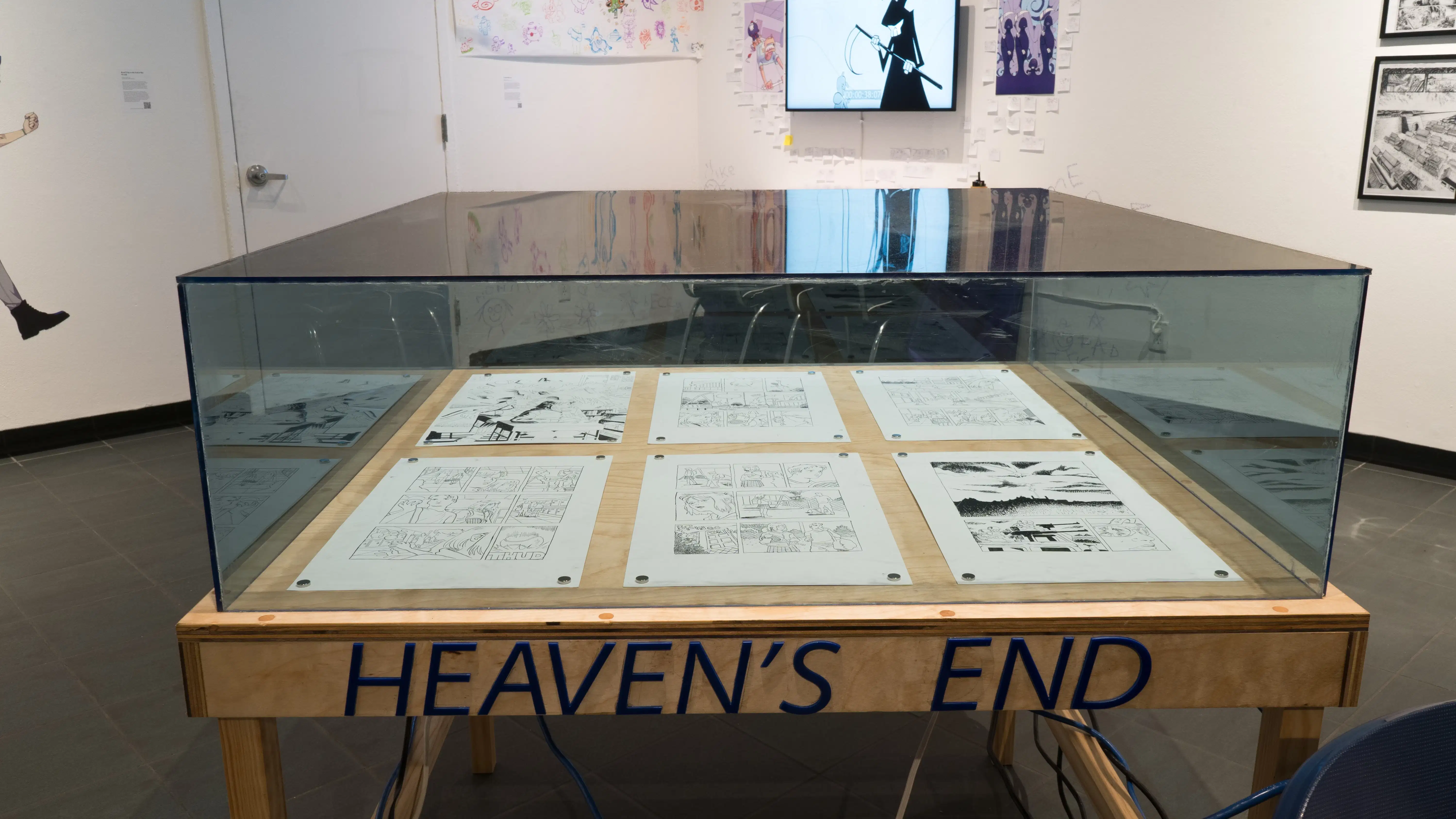 A closer look at the glass display case, showing pages of a comic book resting inside and the words “Heaven’s End” written on the wooden table.