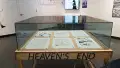 A closer look at the glass display case, showing pages of a comic book resting inside and the words “Heaven’s End” written on the wooden table.