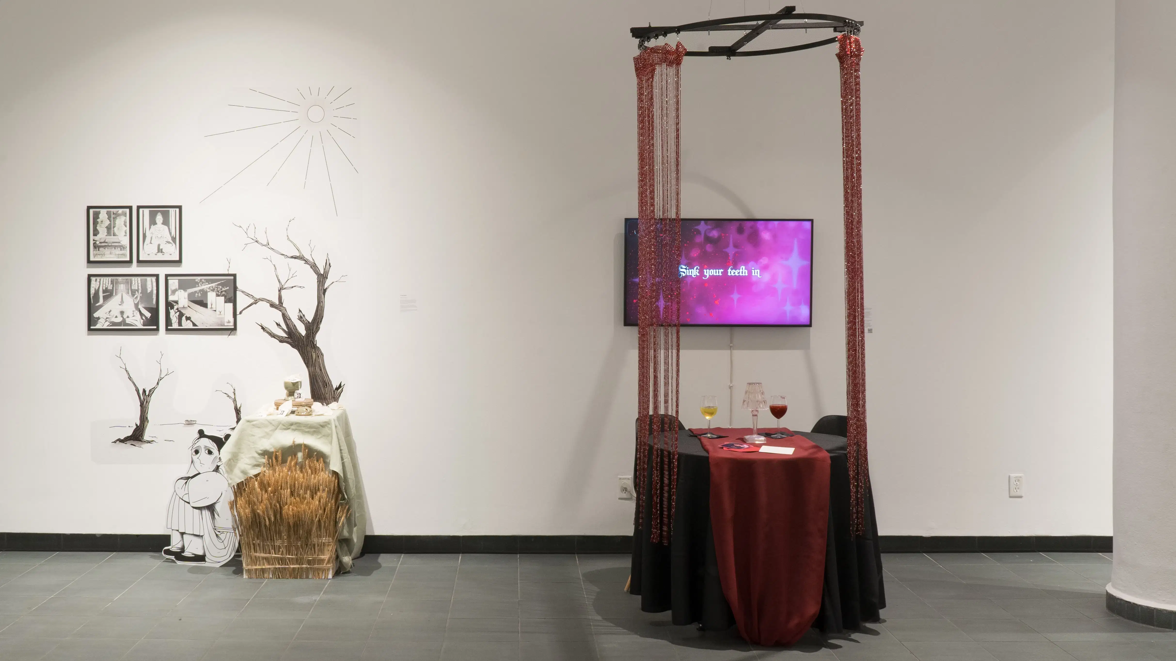 Two works in the exhibition. On the left, there are four black and white illustrations, as well as images of dying trees stuck to the wall, and a green tablecloth over some wheat. To the right of that is an animation. In front of the animation is a small, round table with two wine glasses and two chairs, which is surrounded by glittery tassels that hang from above the television.