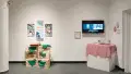 Two works in the exhibition. On the left, four artworks hang above a series of crates. A chessboard balances on top of the crates. On the right, a video plays on a television . Several works of embroidery hang on the walls beside the video, and a plinth sits below with a tank on top of it.