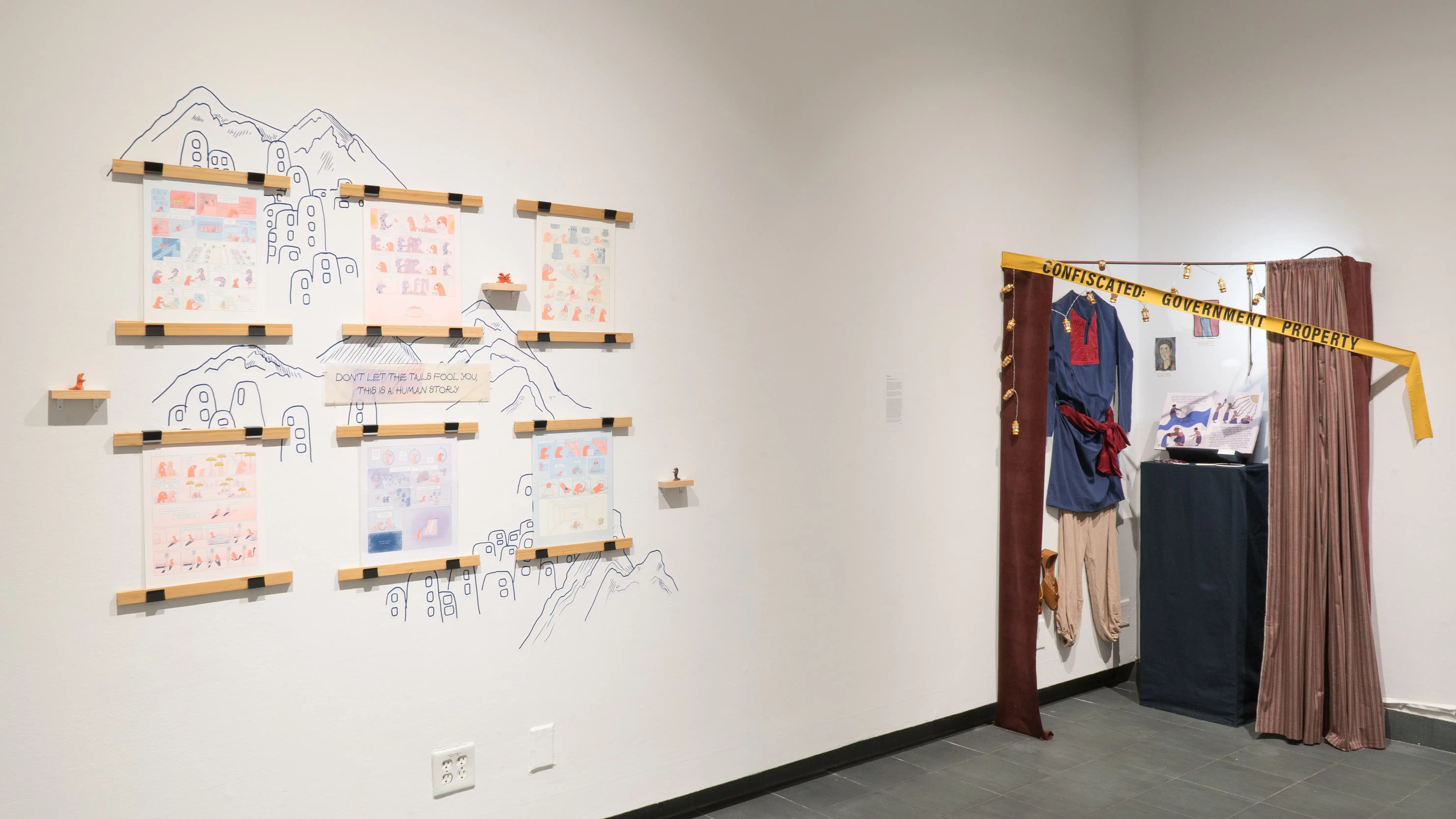 Two works in the exhibition. On the left, a colourful series of comic pages hang on the wall, behind them are mountains represented through blue linework. A plaque reading “Don’t let the tails fool you, this is a human story” sits centred in the piece. To the right of that is a series of items in a corner of the gallery. Clothes and shoes hang on the wall, close to a plinth that has an open book on top of it. The items are behind curtains that are mostly drawn, and yellow tape that reads “Confiscated: Government Property.”
