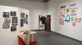 11 framed artworks on a divider in the middle of the room. A small autumn themed table is in front of the works. In the corner of the room is an outfit hung on the wall, partially hidden behind a curtain. To the right is an animation surrounded by graphic stickers, also made by the artist.