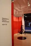 Installation shot showing a black plexiglass organic shape hanging from the middle of the room surrounded by a red curtain and a black plexi circle immediately beneath it.