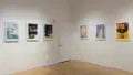 Six poster illustrations hang in a horizontal line along another corner wall in the gallery space, separated by a door. 