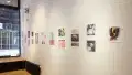 Colorful illustrative prints hang in a horizontal arrangement along the gallery wall next to a large window with a view of the street.  