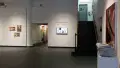 The title wall at the SVA Gramercy Gallery displays wall vinyl and a monitor reading “2024 Thesis Exhibition / Masters in Digital Photography / In Flux.” Closer to the camera, newspapers are stacked on a pedestal and a large photograph printed on a canvas is hung on the wall.