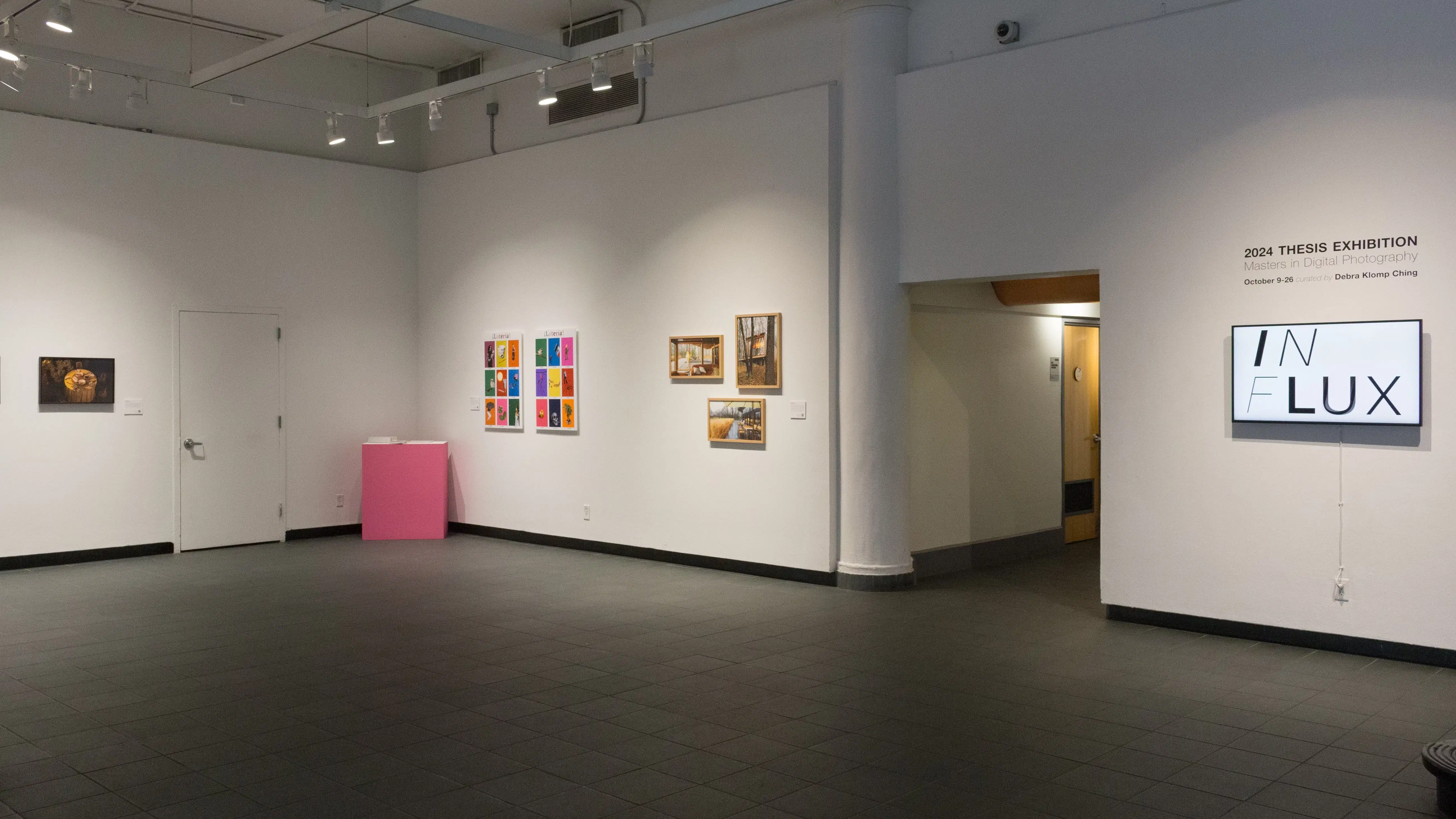 In the lobby gallery, the title wall is visible as well as a few of the works in the exhibition. In the corner, a pink pedestal sits next to two colorful prints. Framed photos containing neutral colors hang on either side.