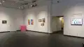 In the lobby gallery, the title wall is visible as well as a few of the works in the exhibition. In the corner, a pink pedestal sits next to two colorful prints. Framed photos containing neutral colors hang on either side.